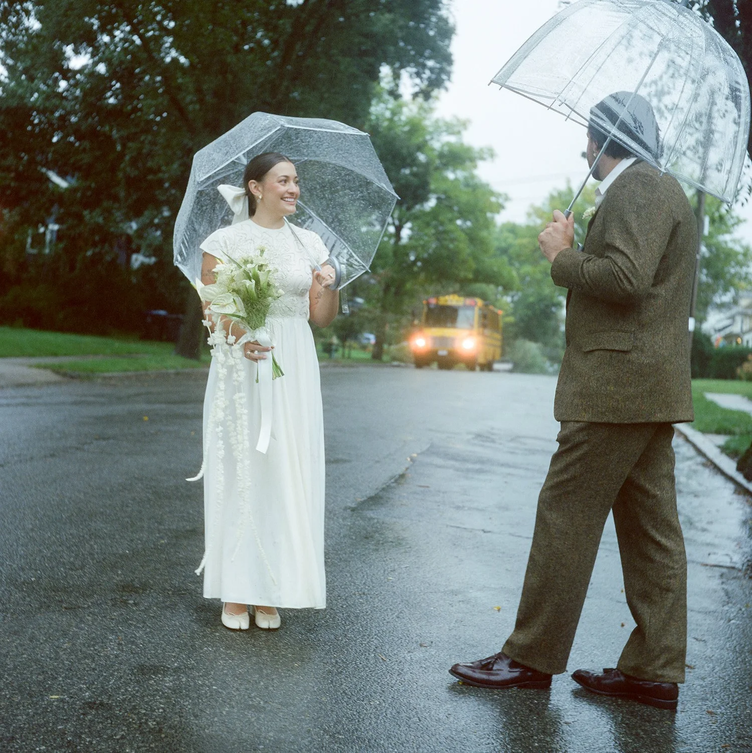 A woman in a white dress holding a bouquet and a man in a suit with umbrellas on a rainy street, trees in background.