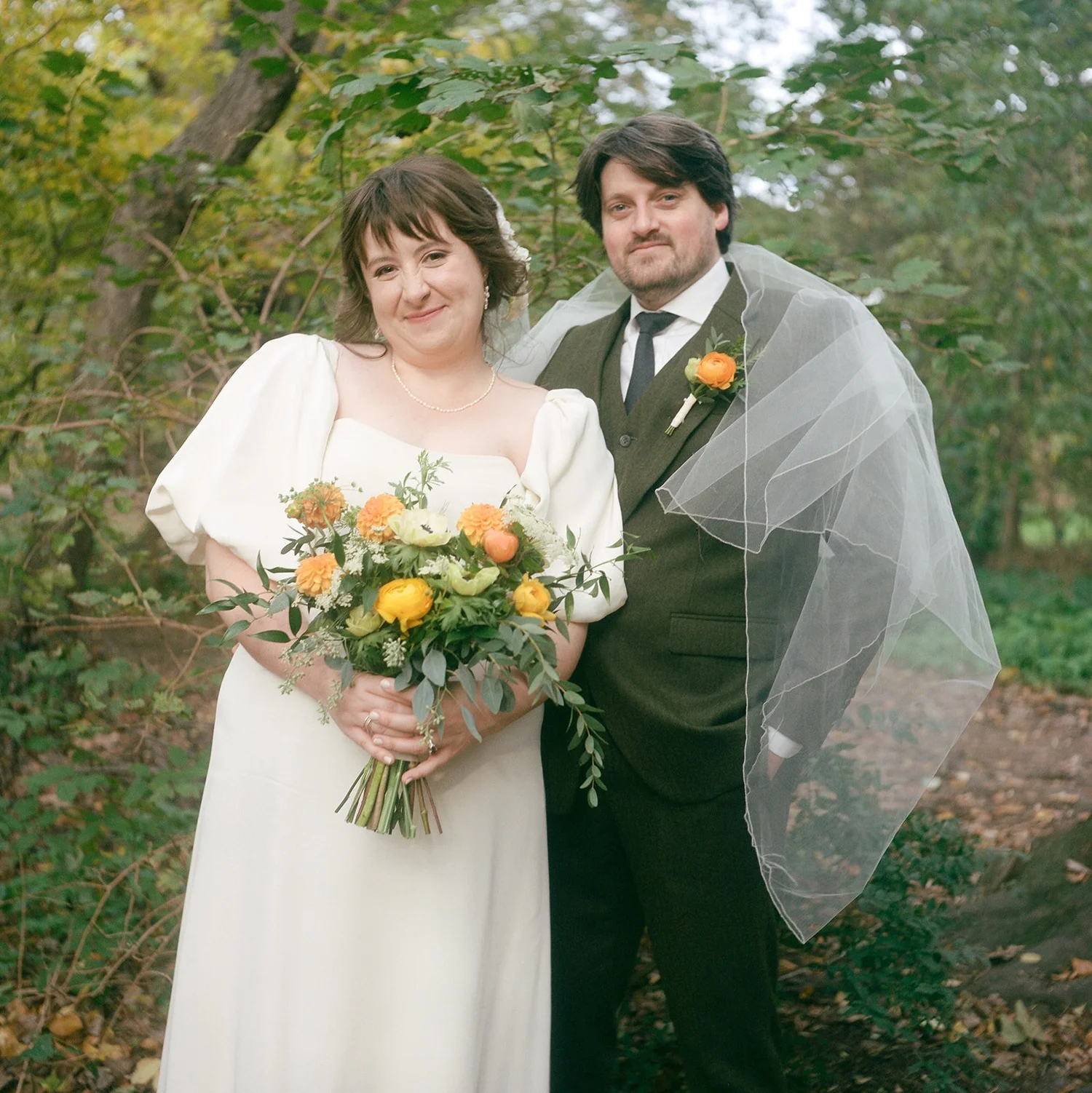 A bride and groom stand outdoors with trees and foliage in the background, smiling, with the bride holding a bouquet of flowers.