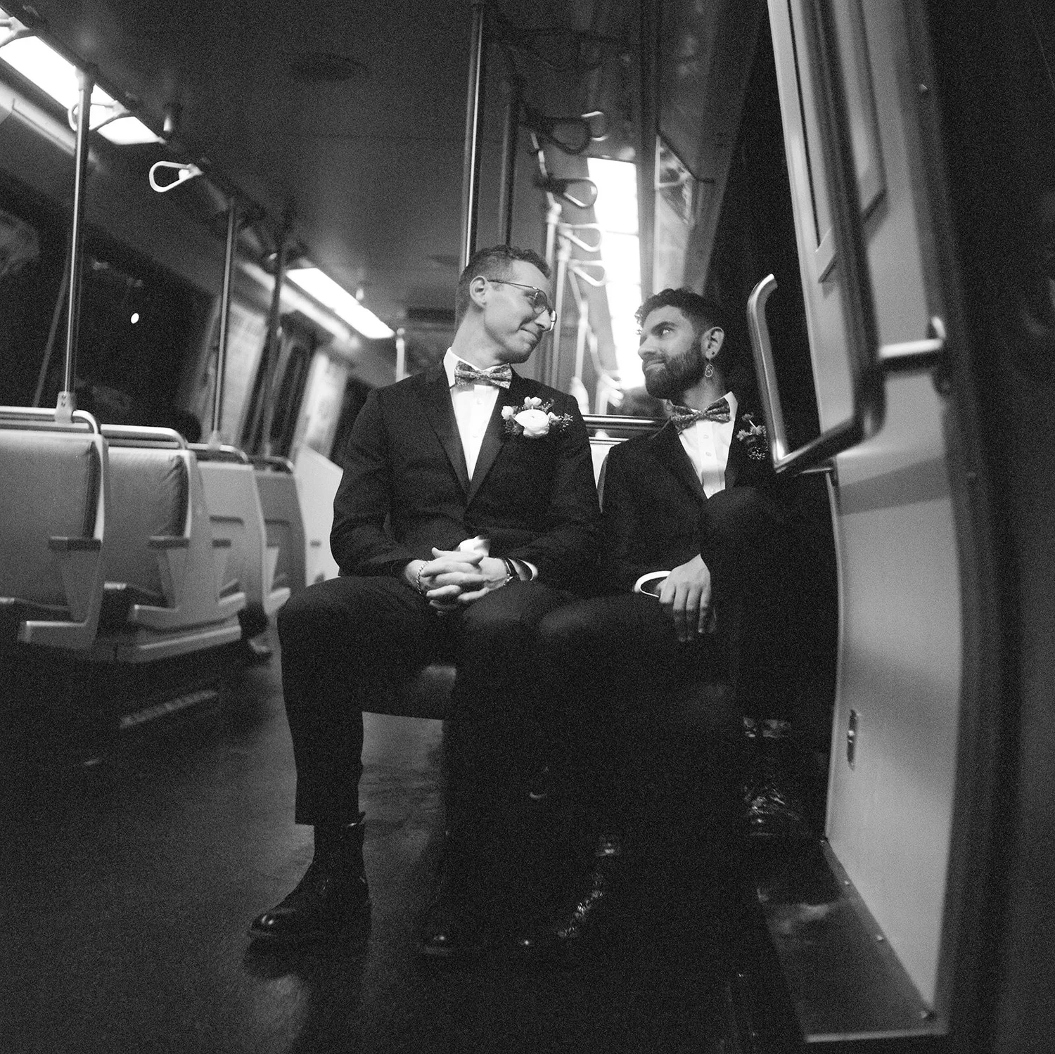 Two men in tuxedos sitting on a bus, facing each other and talking, with empty bus seats visible in the background.