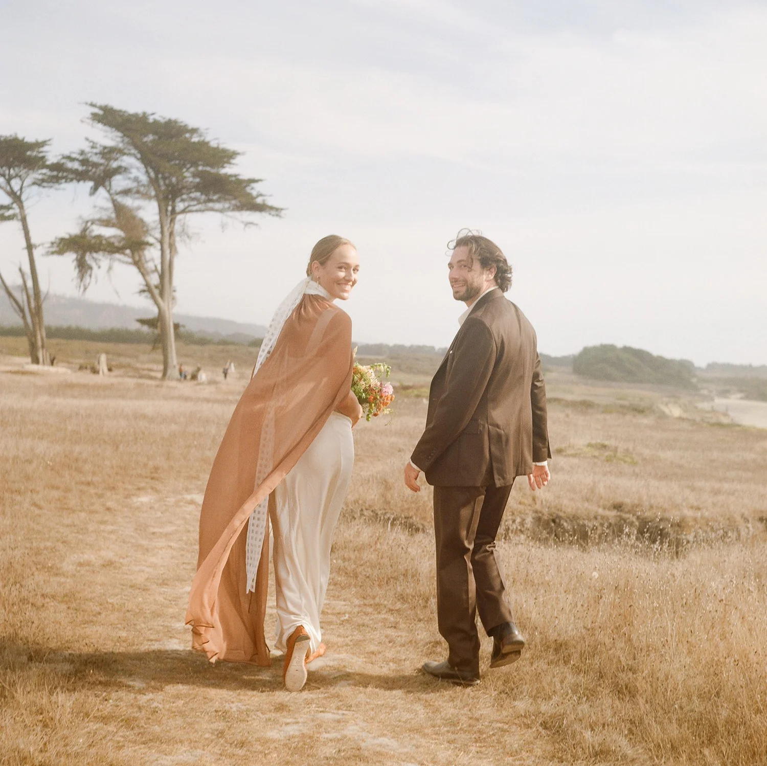 A couple walking outdoors in a field, with trees and a cloudy sky in the background, holding hands and smiling.