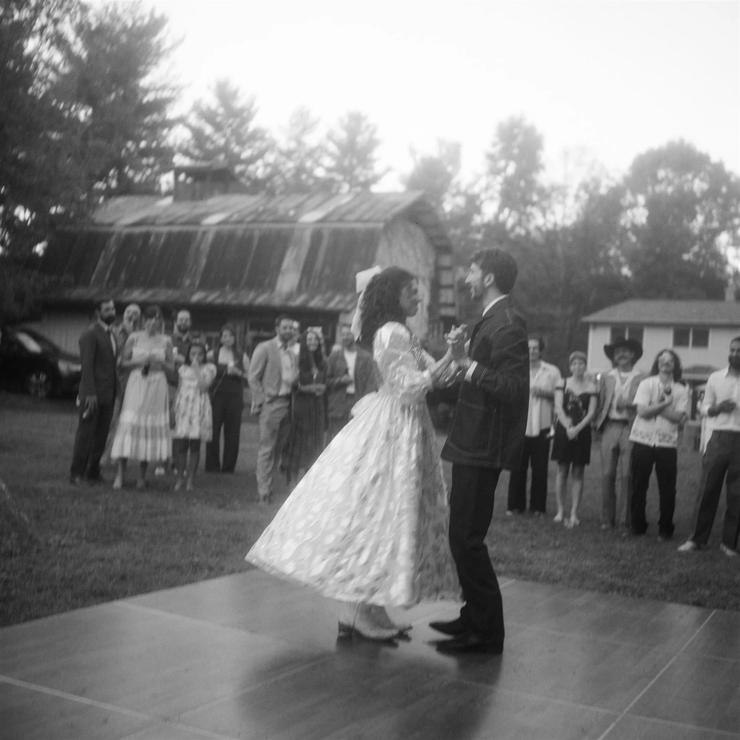 Couple dancing outdoors with a group of people watching, trees, and buildings in the background, black and white photo.