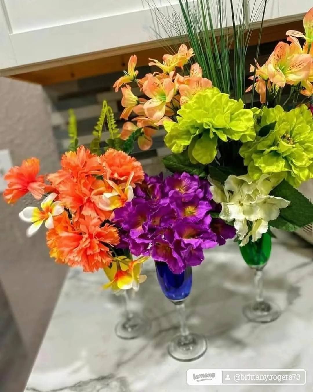 Three colorful flower arrangements in vases on a table, with various flowers including orange, purple, and green, in an indoor setting.