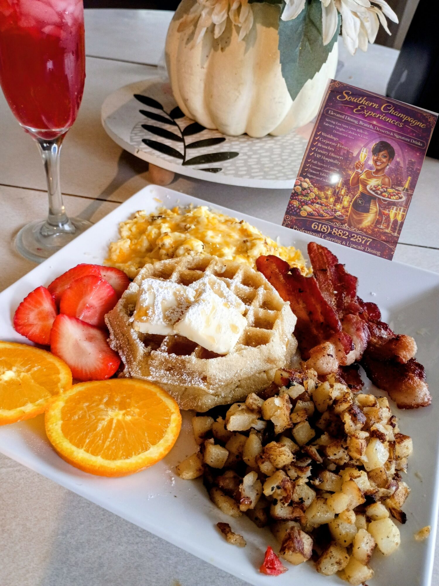 Plate with waffles, bacon, strawberries, orange slices, and scrambled eggs, with a glass of red drink and a pumpkin decoration in background