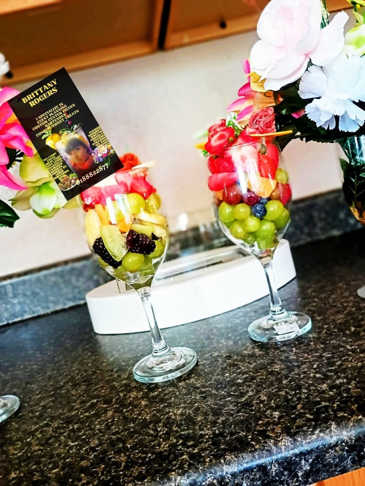 Two tall glasses filled with colorful fruit and candy, decorated with flowers, on a dark countertop.