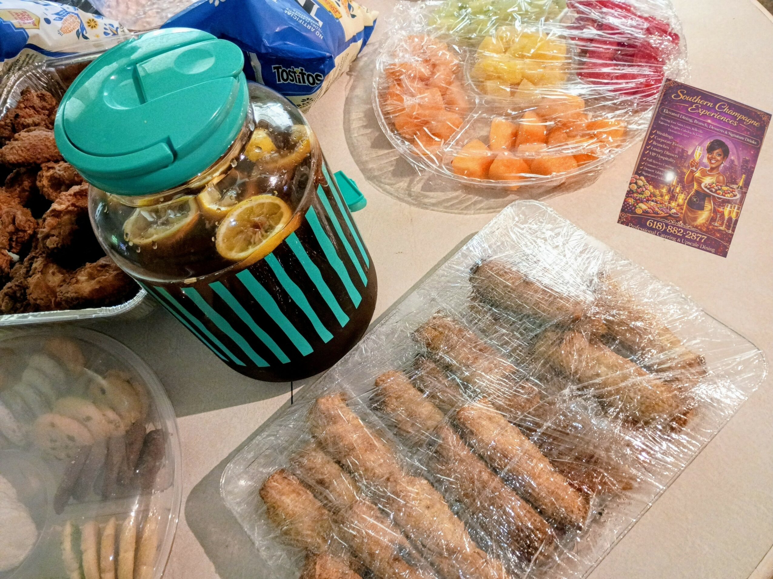 Assorted food items including fried chicken, a jar of pickles, a bowl of fruit, and wrapped snacks on a table.