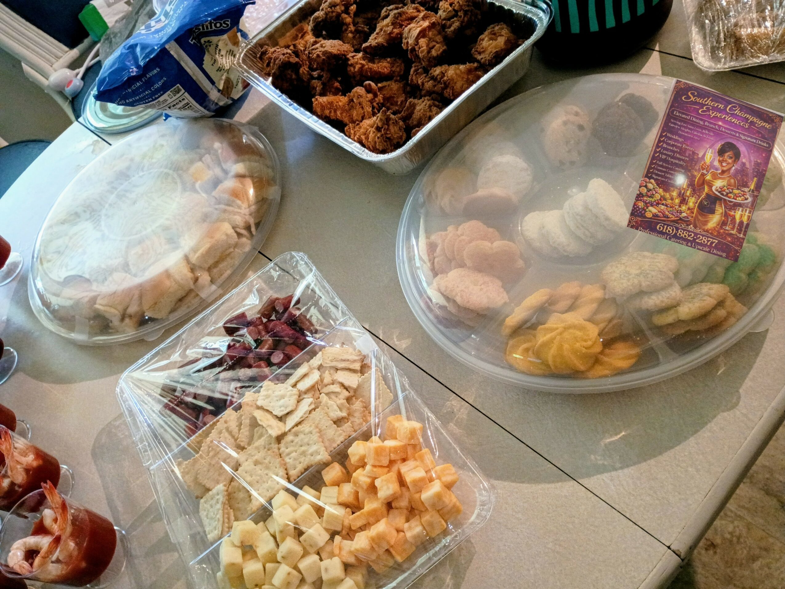 Table with various snacks including fried chicken, cheese and meat cubes, cookies, and a card, viewed from above.