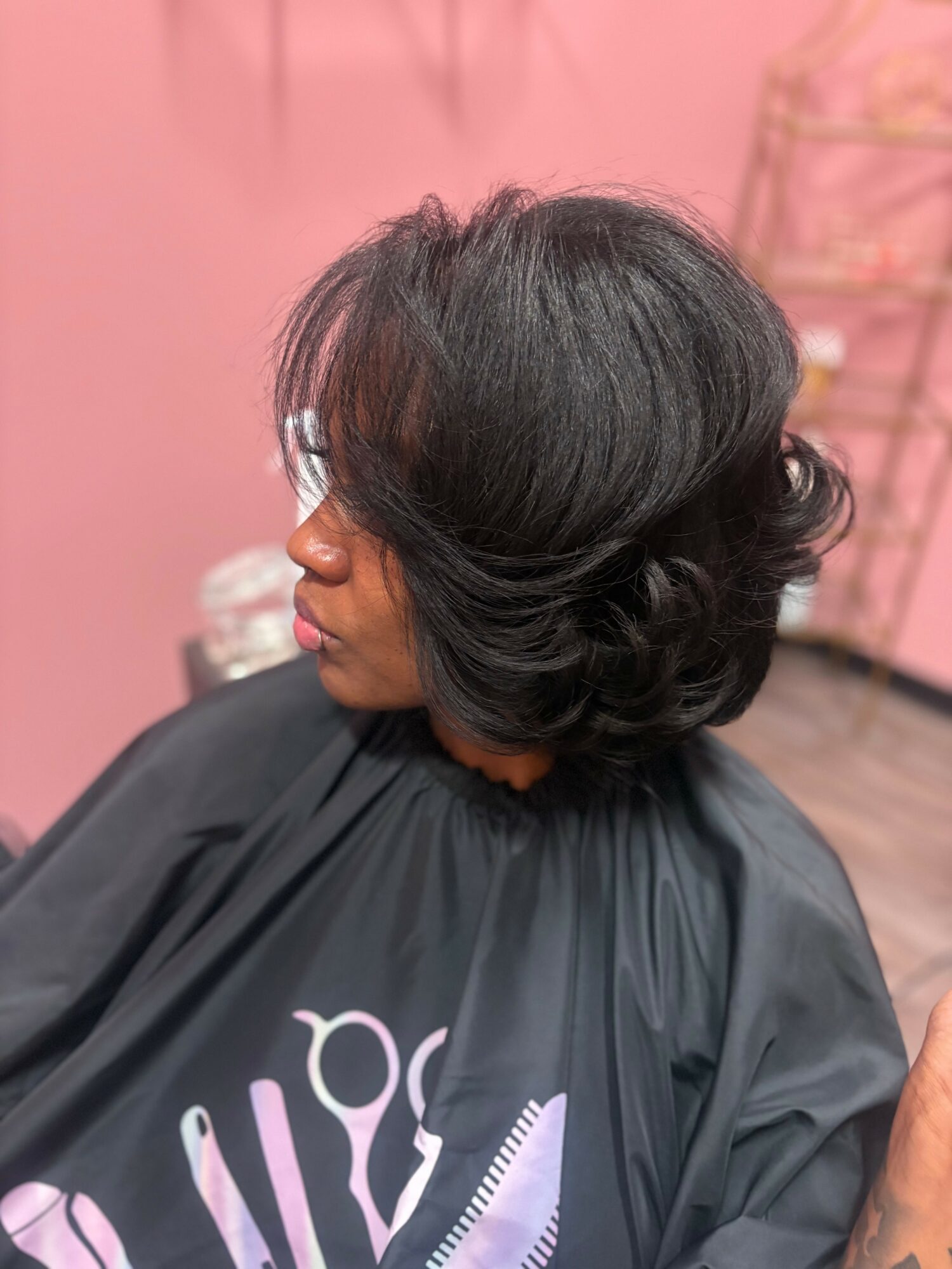 Side view of a woman with a short, layered black hairstyle, sitting in a salon with a pink wall background.