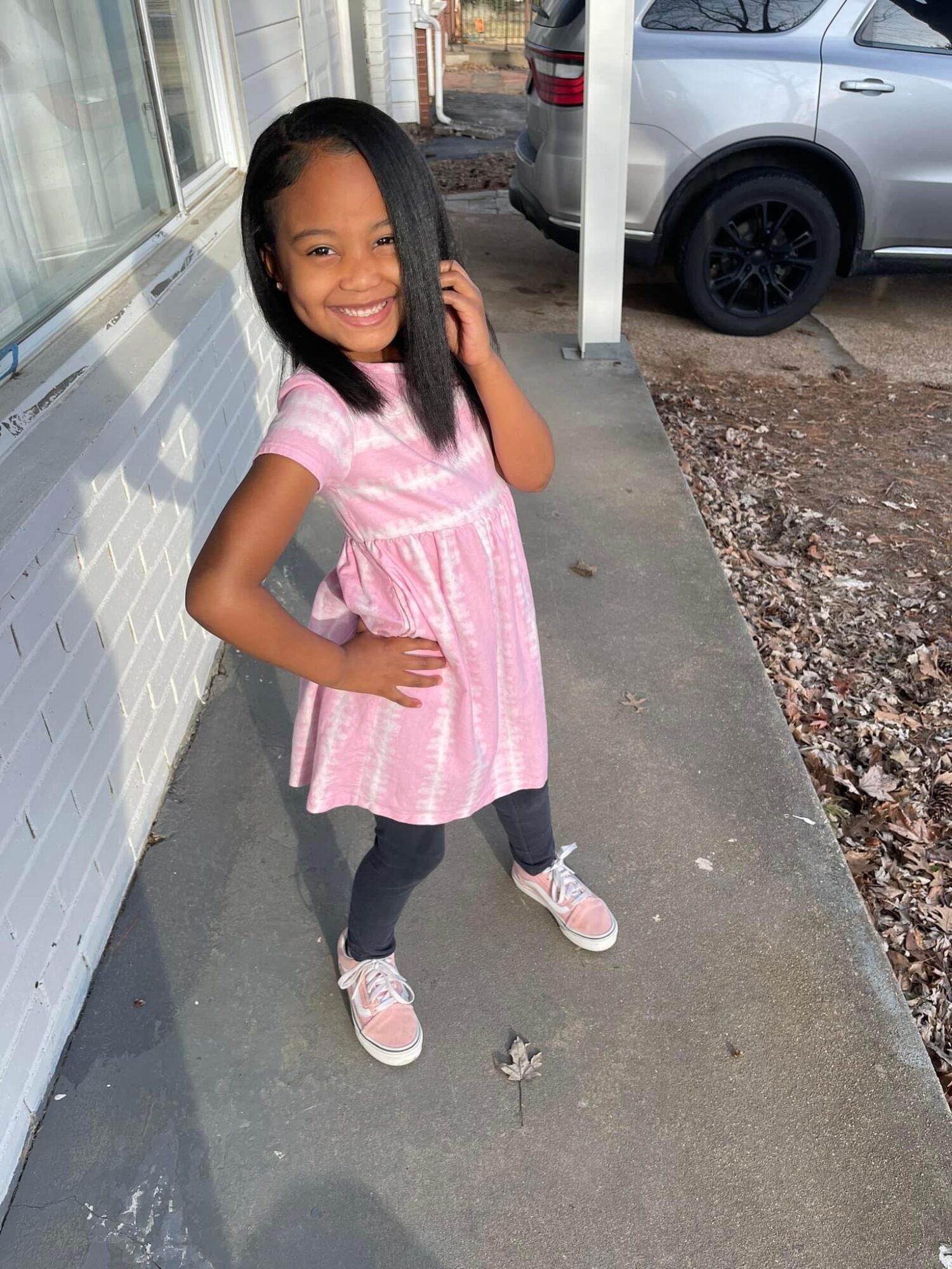 Smiling girl in pink dress and gray leggings standing on sidewalk outside, with a car and building in background.