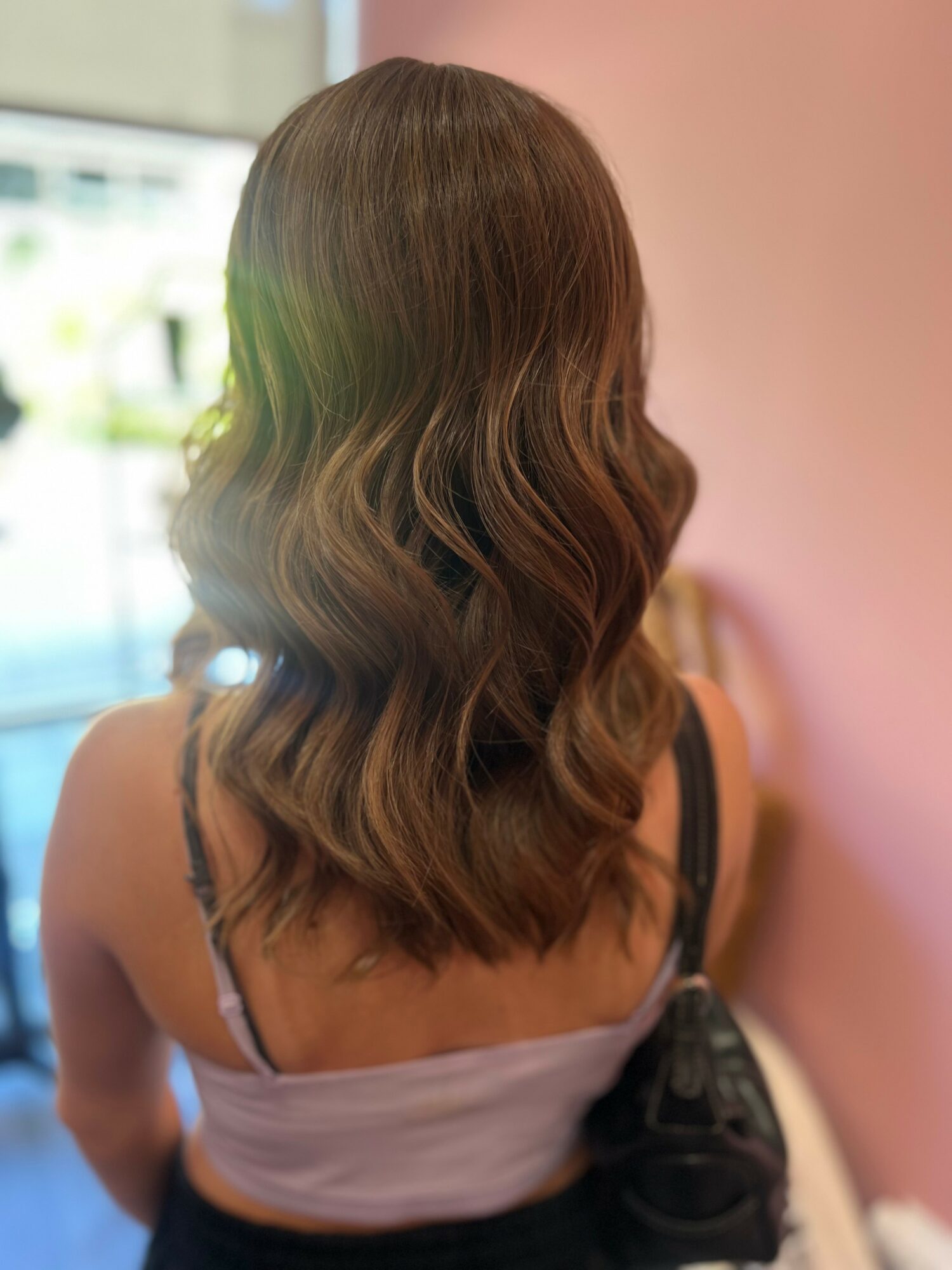 Back view of a woman with wavy, shoulder-length brown hair, wearing a light-colored top and black straps, standing indoors.