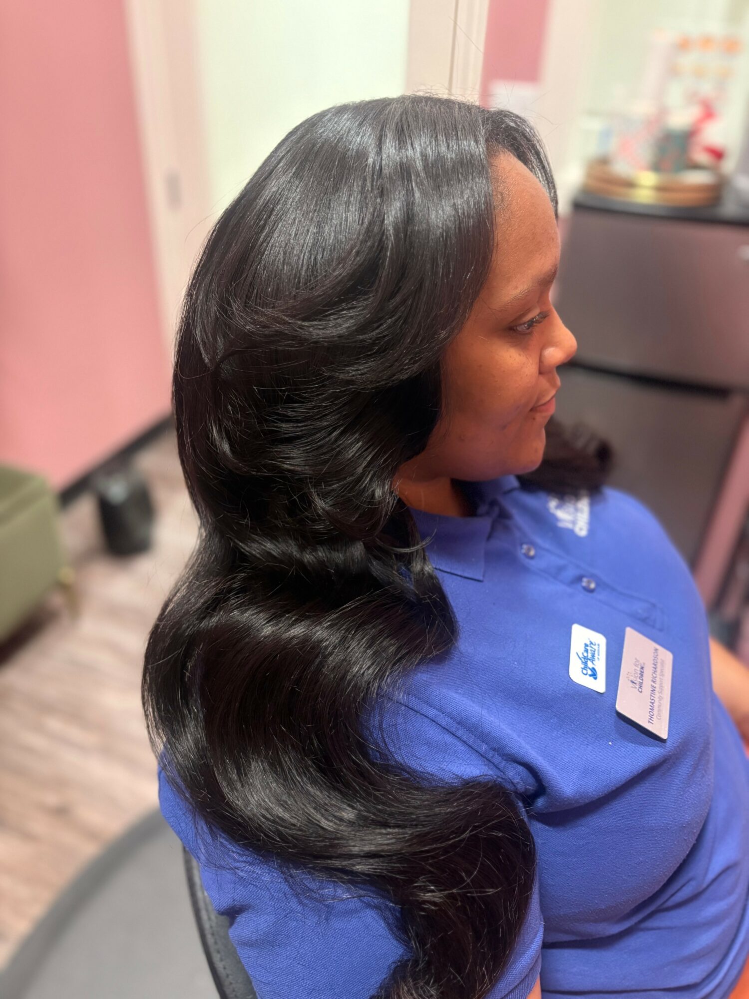 Side view of a woman with long, shiny, wavy black hair wearing a blue shirt, sitting indoors.