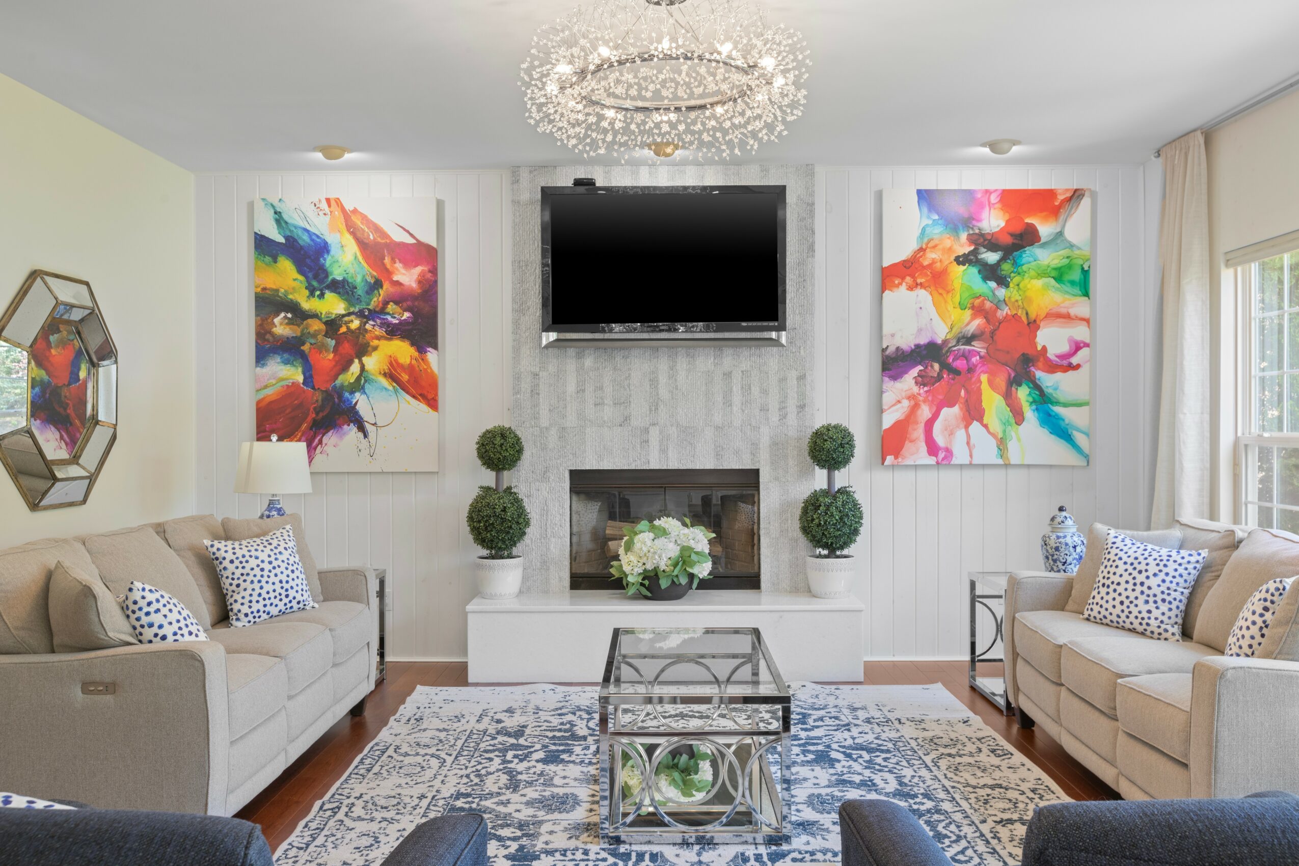 Living room with two sofas, colorful abstract paintings, a chandelier, and a flat-screen TV above a fireplace.