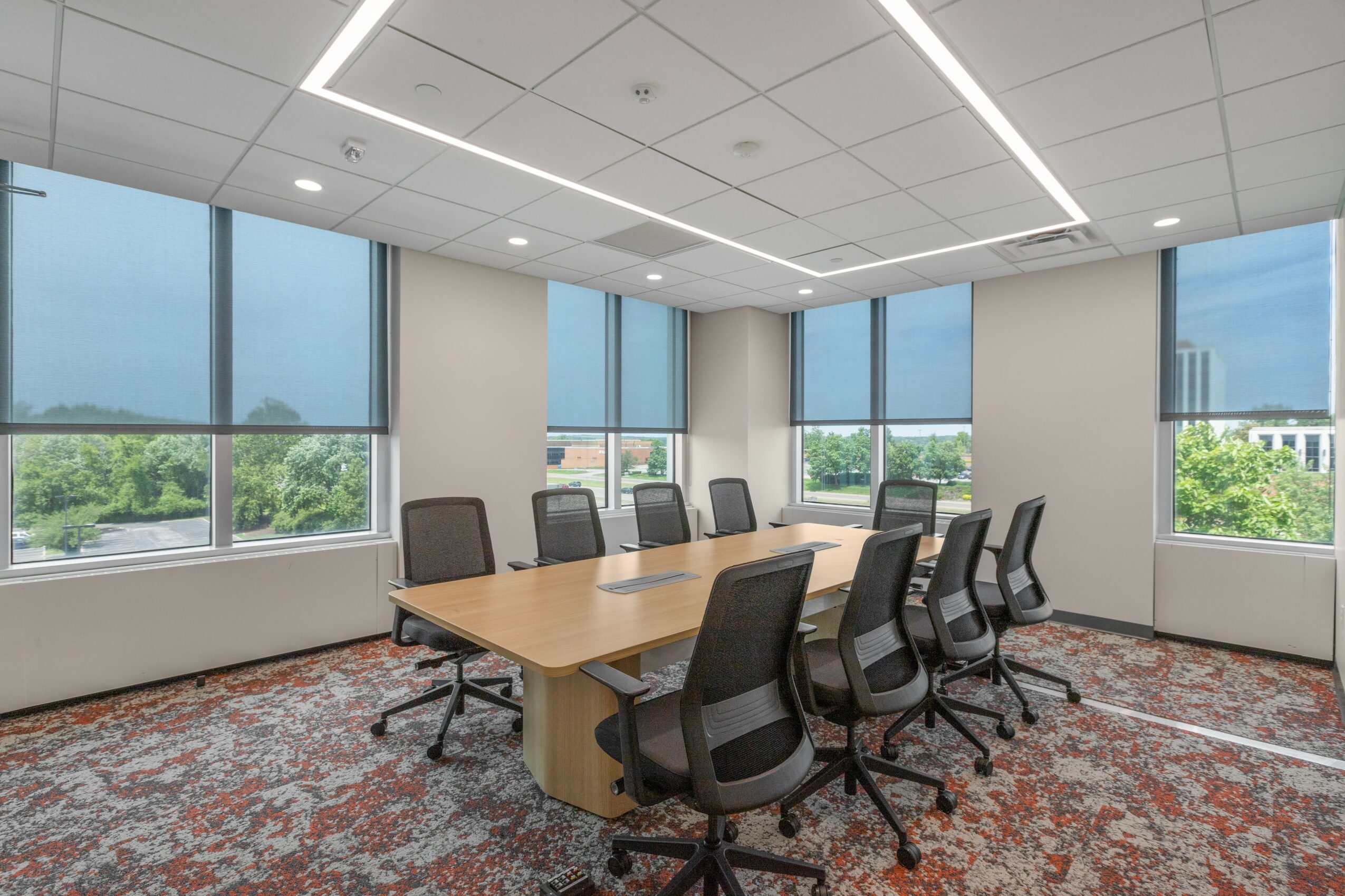 Modern conference room with large windows, a wooden table, and black office chairs, with a patterned carpet and ceiling lights.