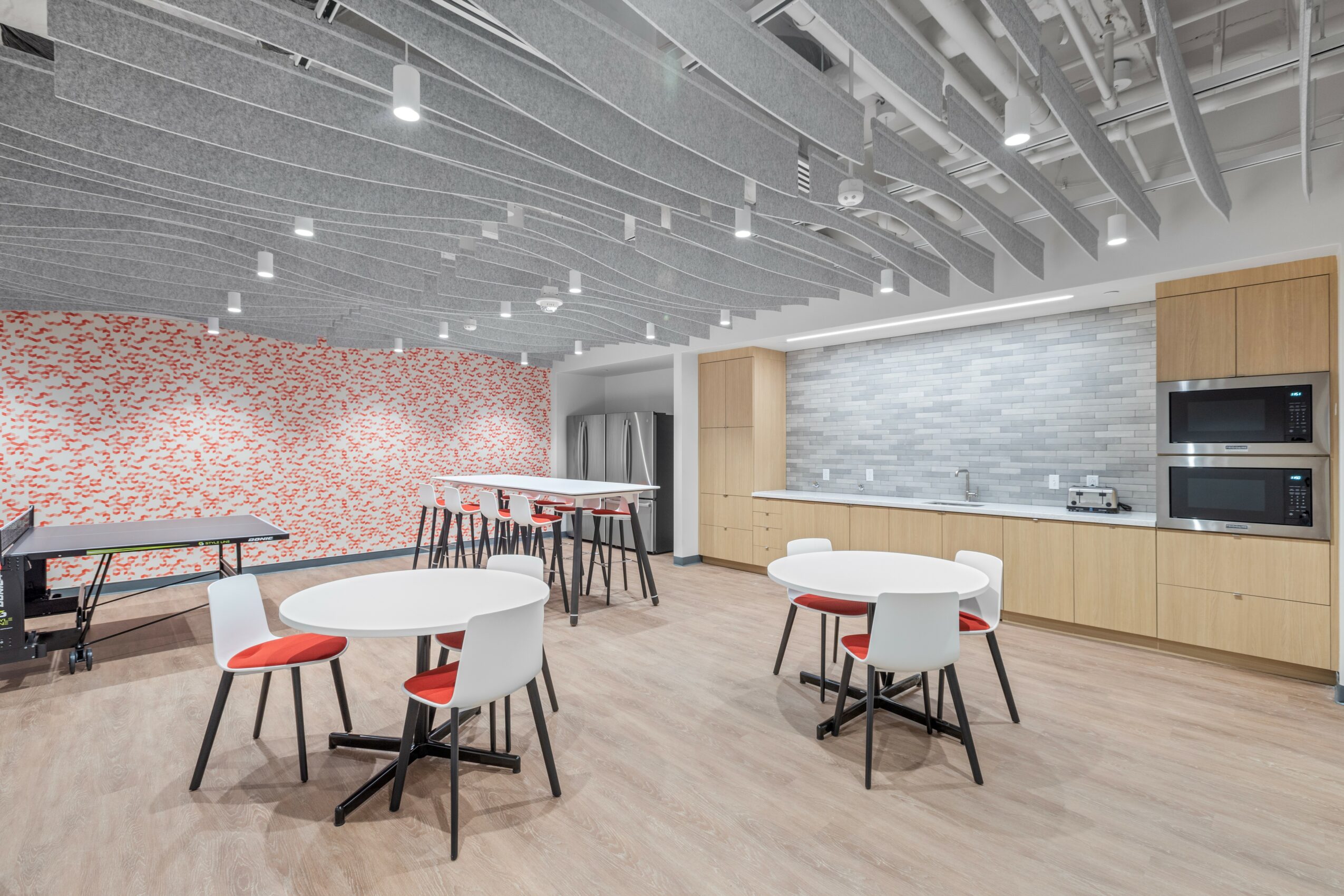 Modern kitchen and dining area with tables, chairs, a pink floral wall, and a ceiling with exposed pipes and lighting.