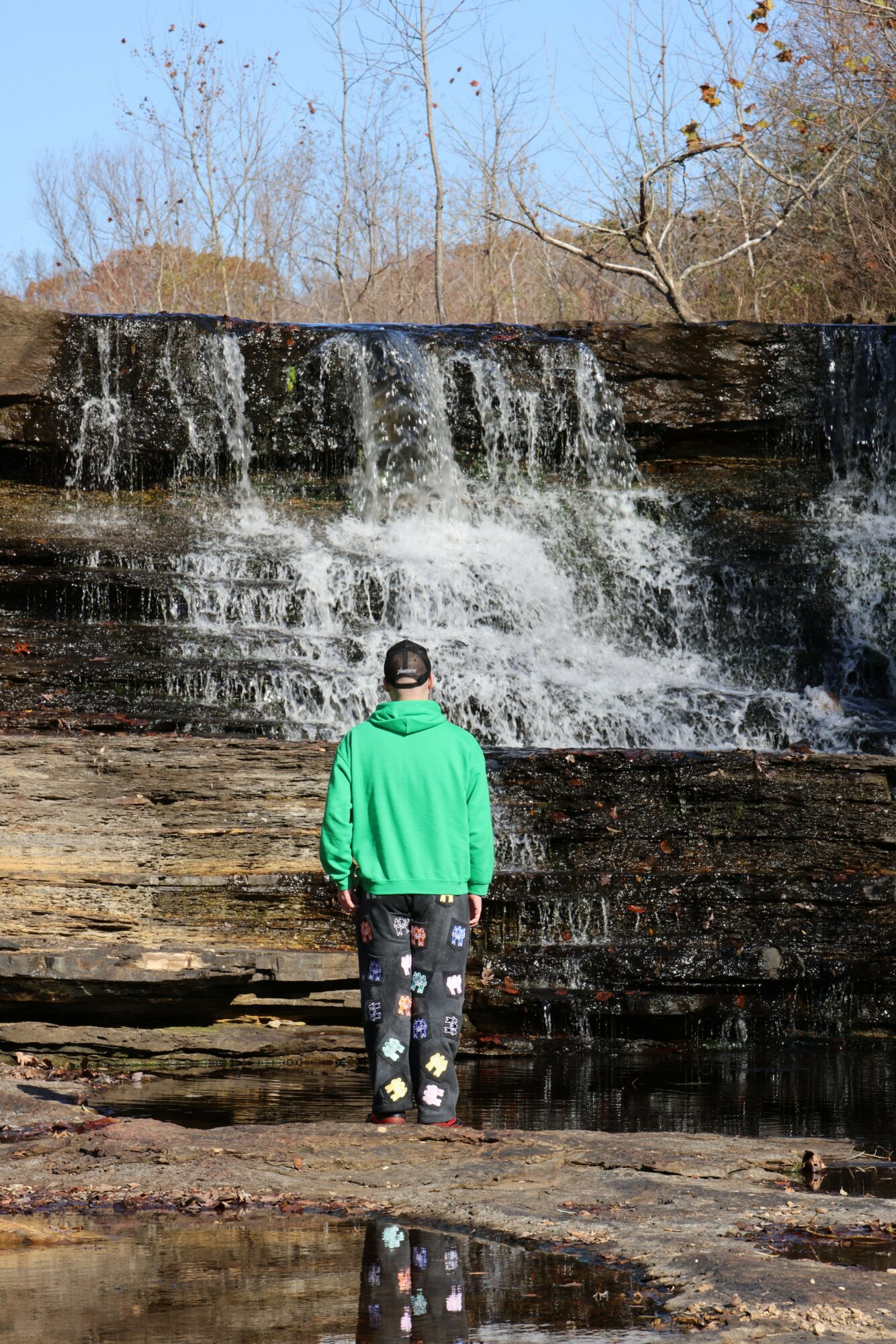 Person standing on rocks near a waterfall in a natural setting, with trees and blue sky in the background.
