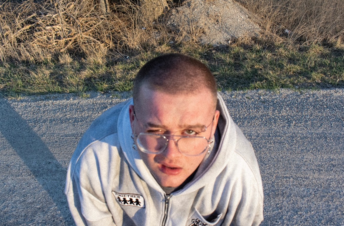 Young person with glasses looking up, wearing a light-colored hoodie, outdoors on a paved surface with grass and bushes behind.