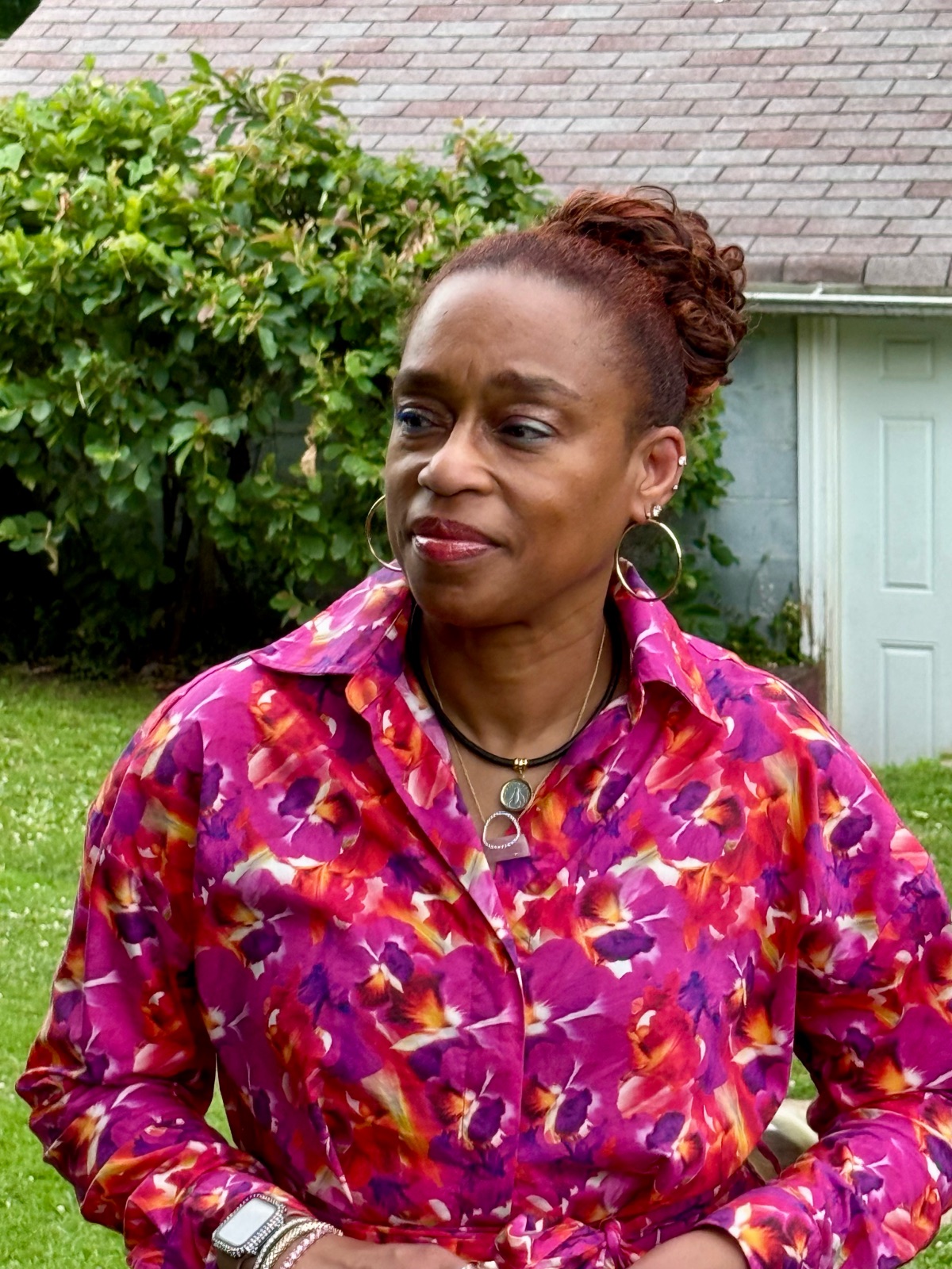 Woman with short curly hair wearing a colorful floral blouse outdoors, with greenery and a building in the background.
