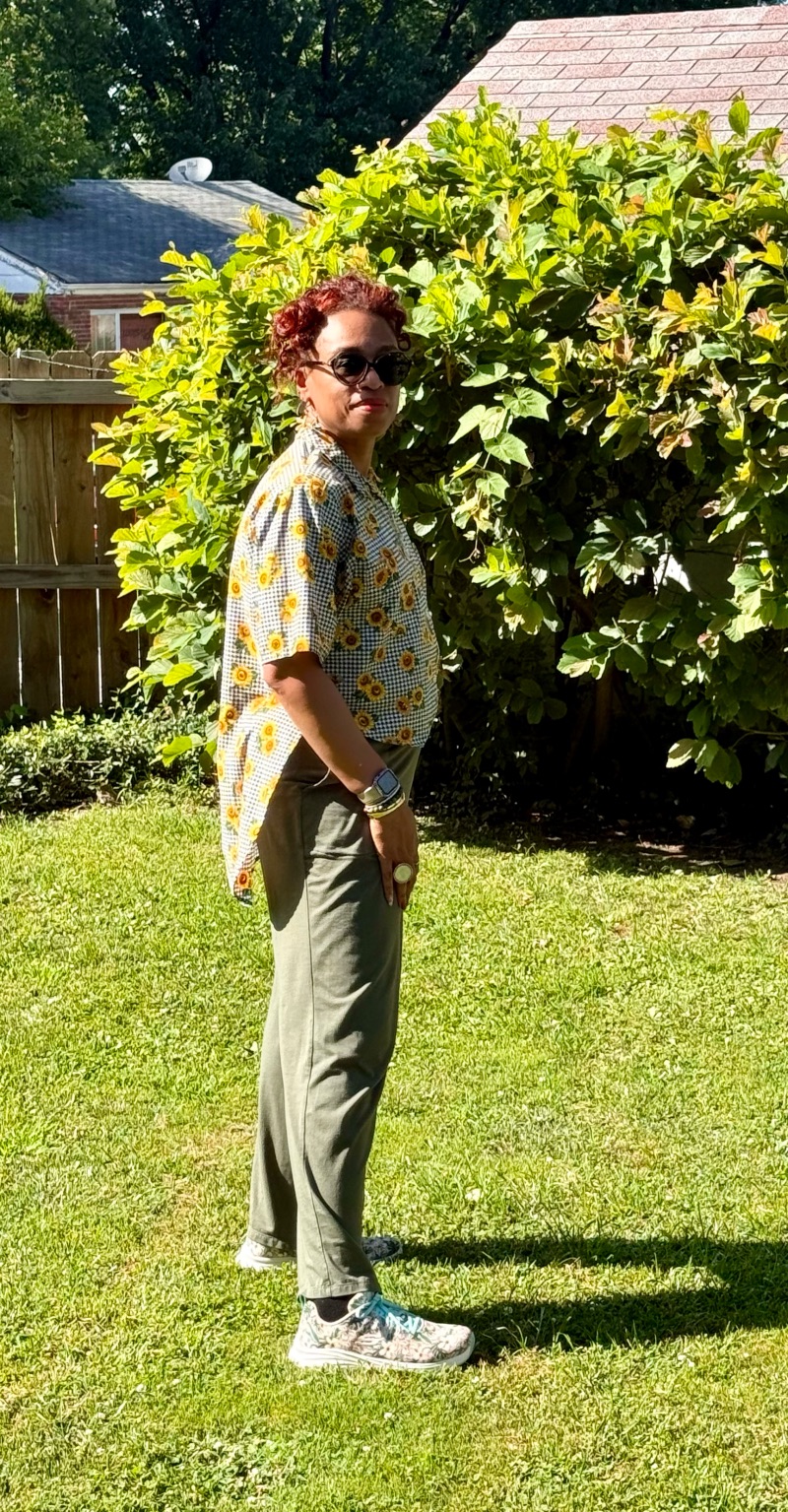 Person standing outdoors on grass with a large leafy bush behind, wearing sunglasses, patterned shirt, and gray pants.