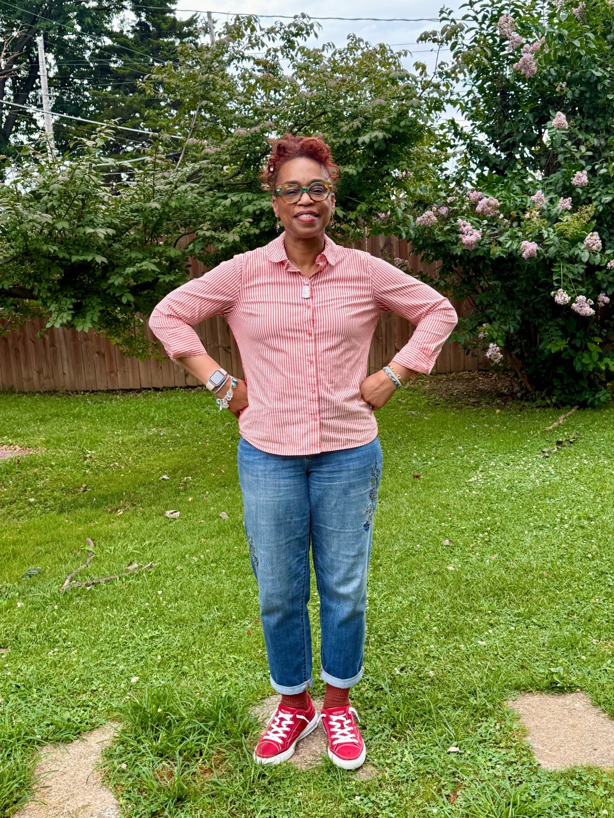 Woman standing outdoors on grass, smiling, wearing pink shirt, blue jeans, and red sneakers, with trees and flowering bushes in background.