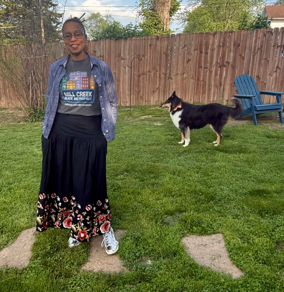 Person standing on grass with a dog in backyard, wooden fence and blue chair in background.