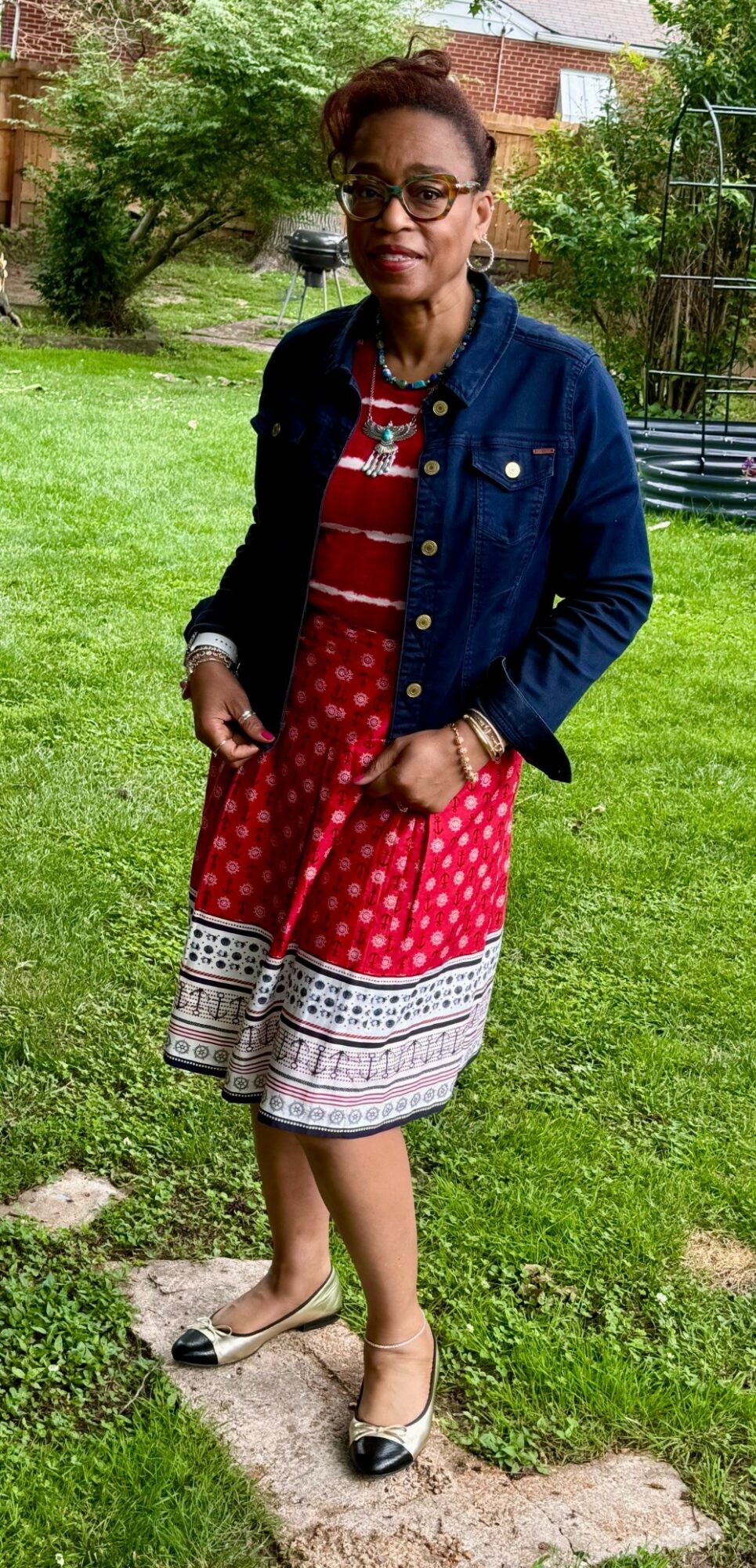 Woman standing outdoors on grass, wearing glasses, a denim jacket, and a red patterned dress, with trees and a fence in background.