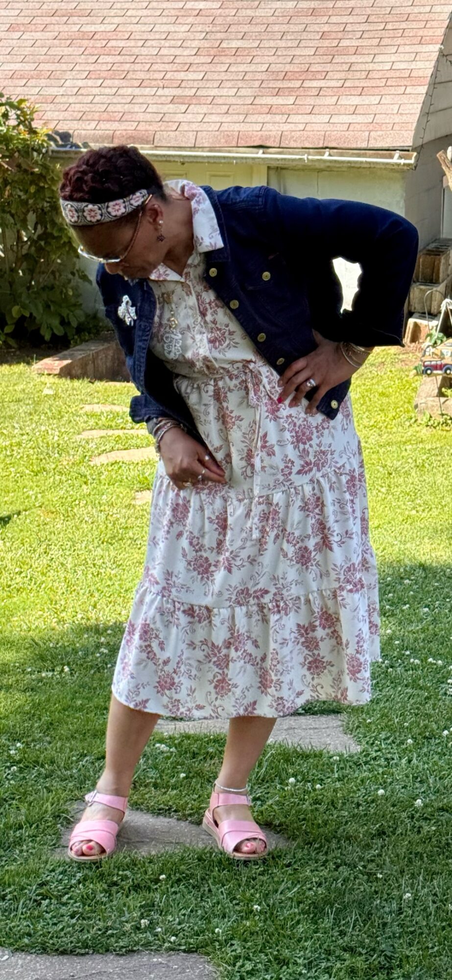 Person bending over outdoors on grass, wearing a floral dress, denim jacket, pink shoes, and headband.