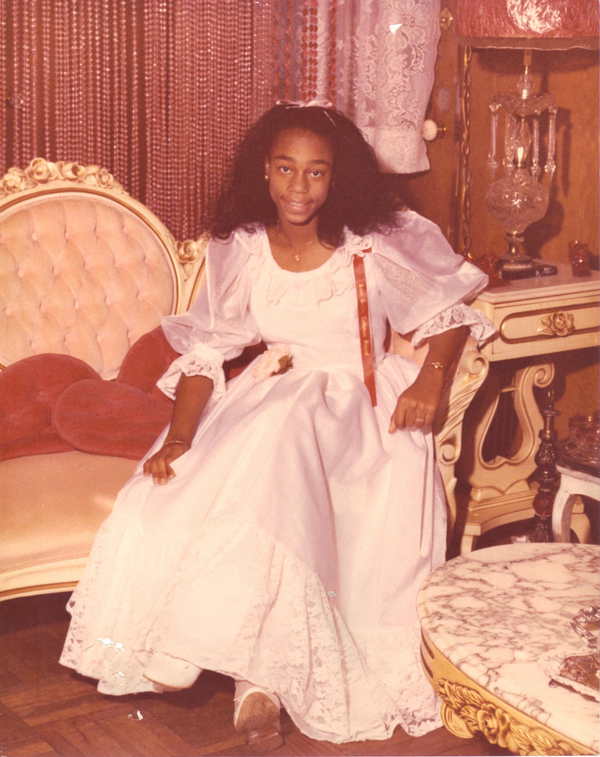 Young girl in a white dress standing in a decorated room with vintage furniture and curtains.
