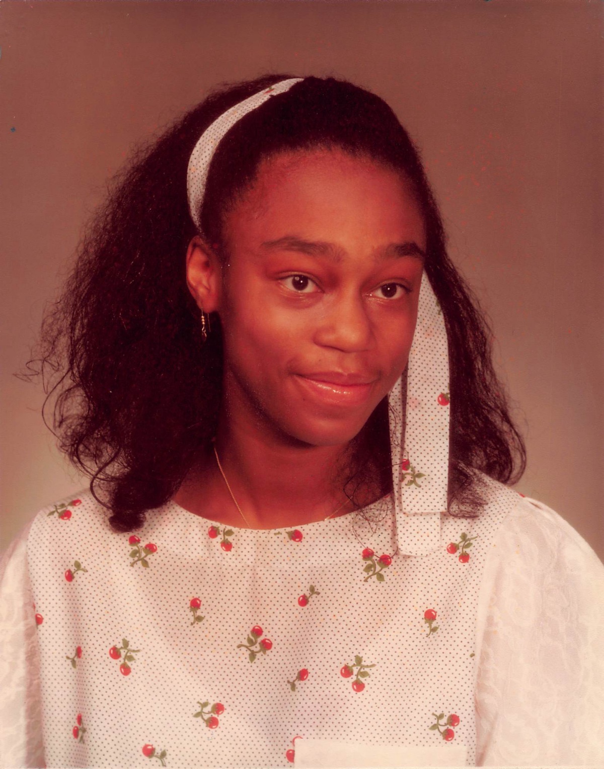 Young girl with curly hair, wearing a white headband and a white dress with small red and green floral patterns.