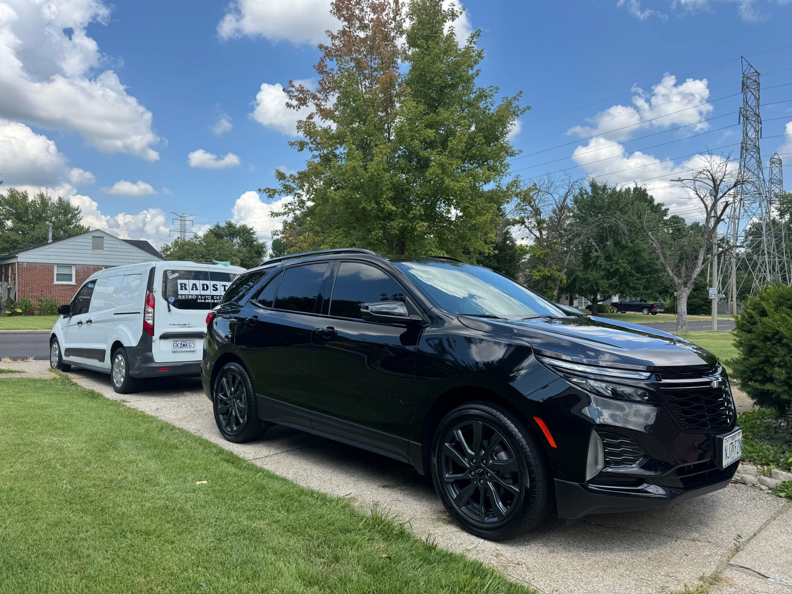 Two parked cars, one black and one white, on a driveway with trees and a house in the background under a blue sky.