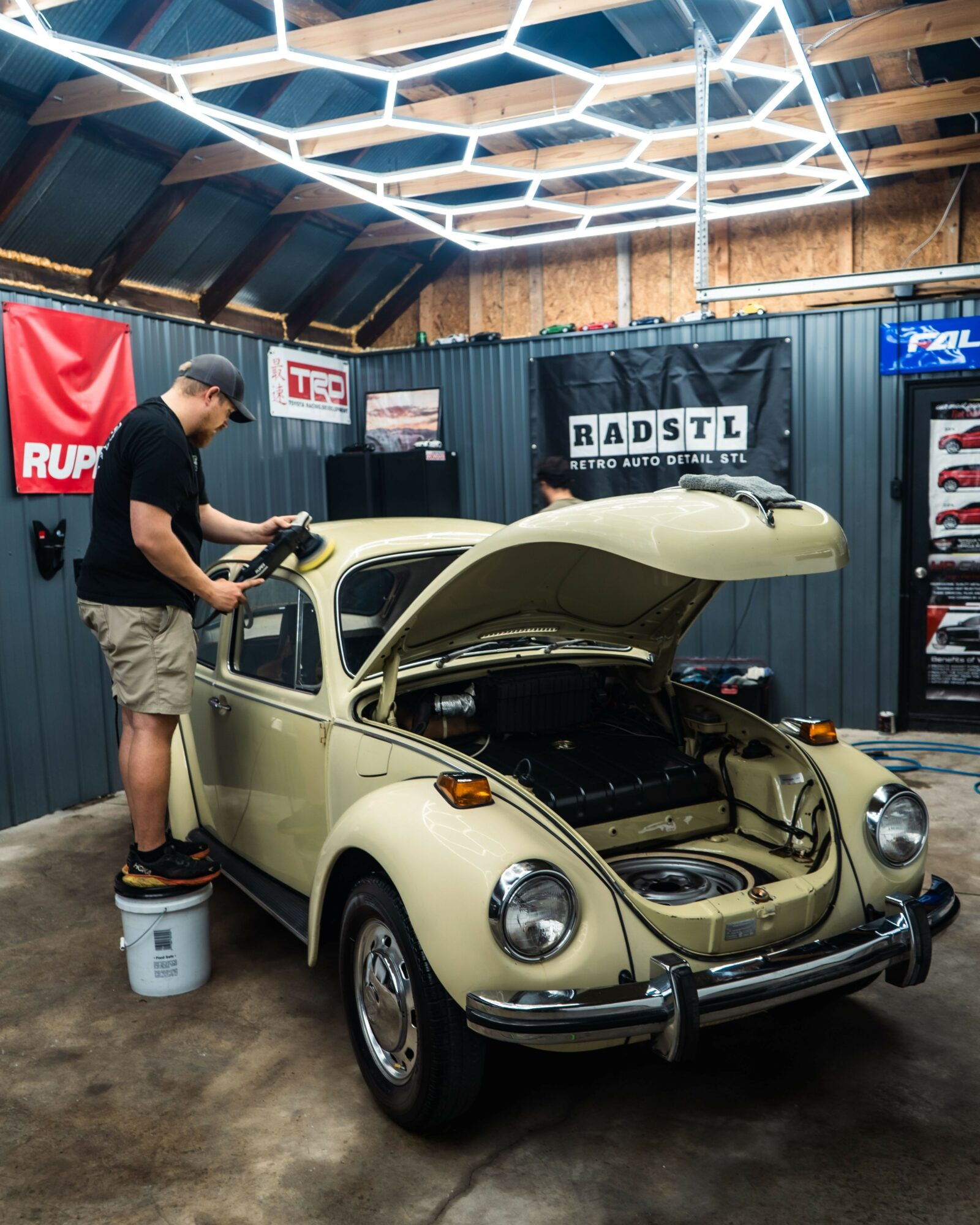Man working on a vintage beige car with open hood inside a garage, with banners and shelves in background.