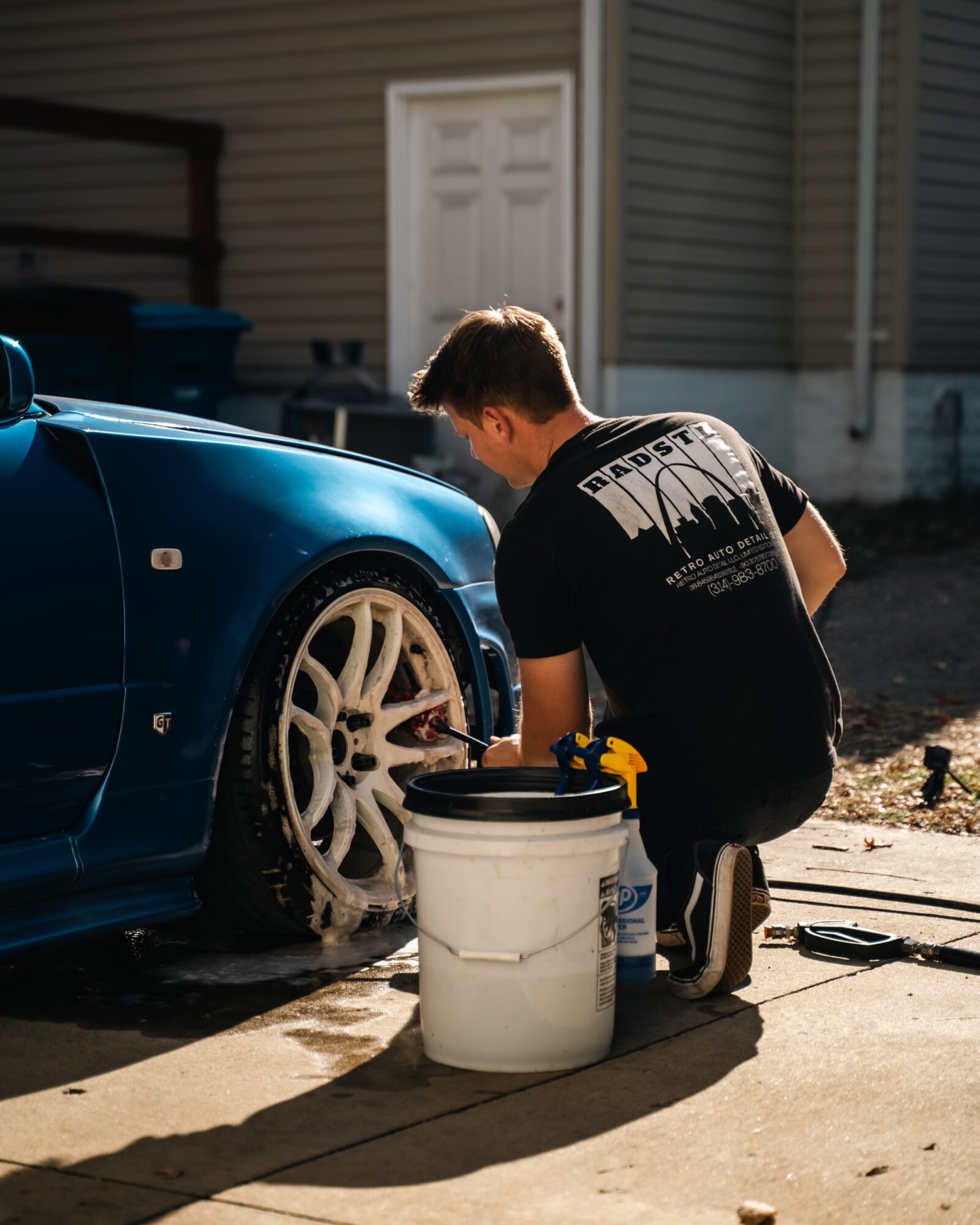 Person cleaning a blue car's wheel outside a house, with a white bucket nearby, sunlight casting shadows.