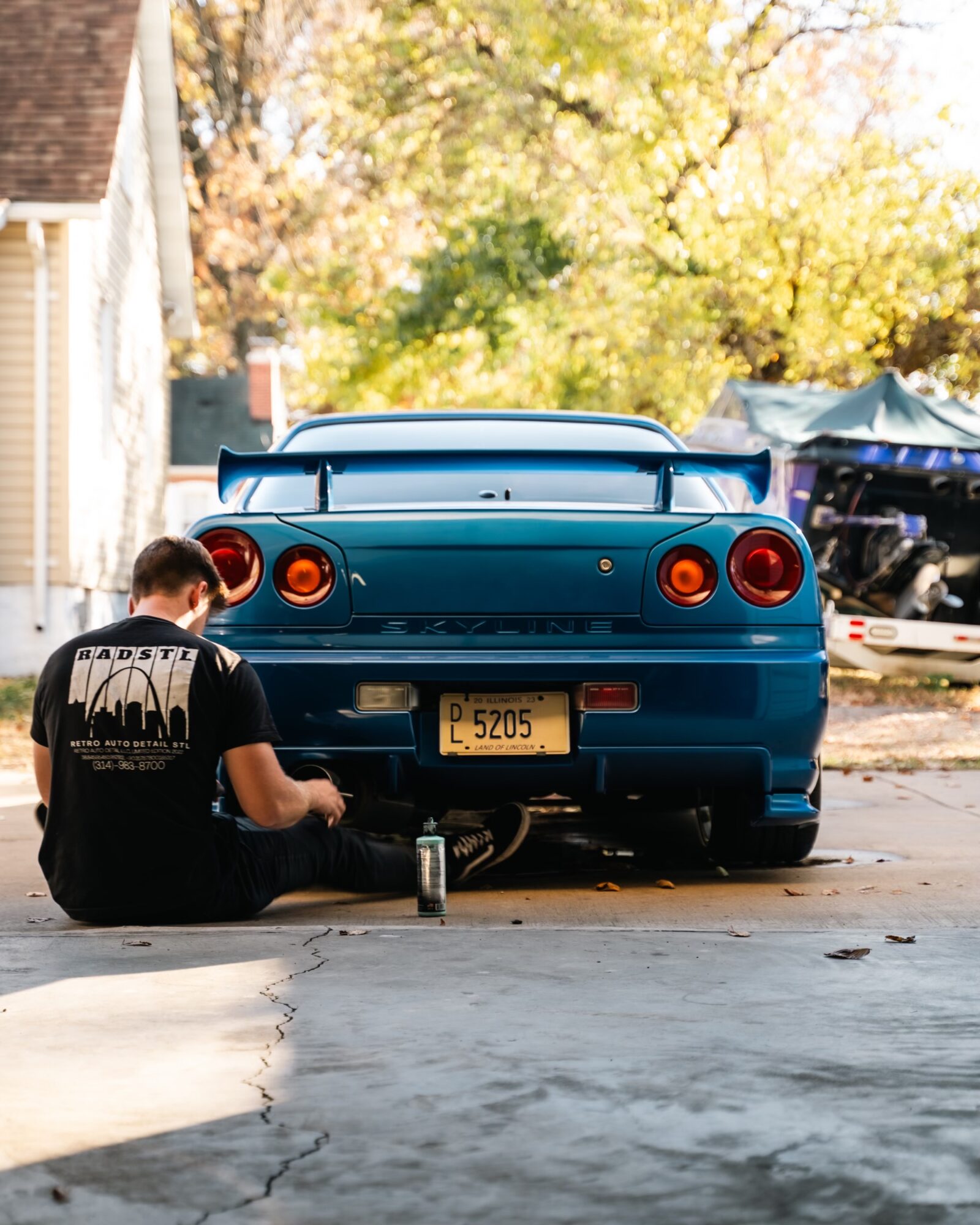 Person sitting on the ground next to a blue sports car with a yellow license plate, outdoors with trees and a tent in background.