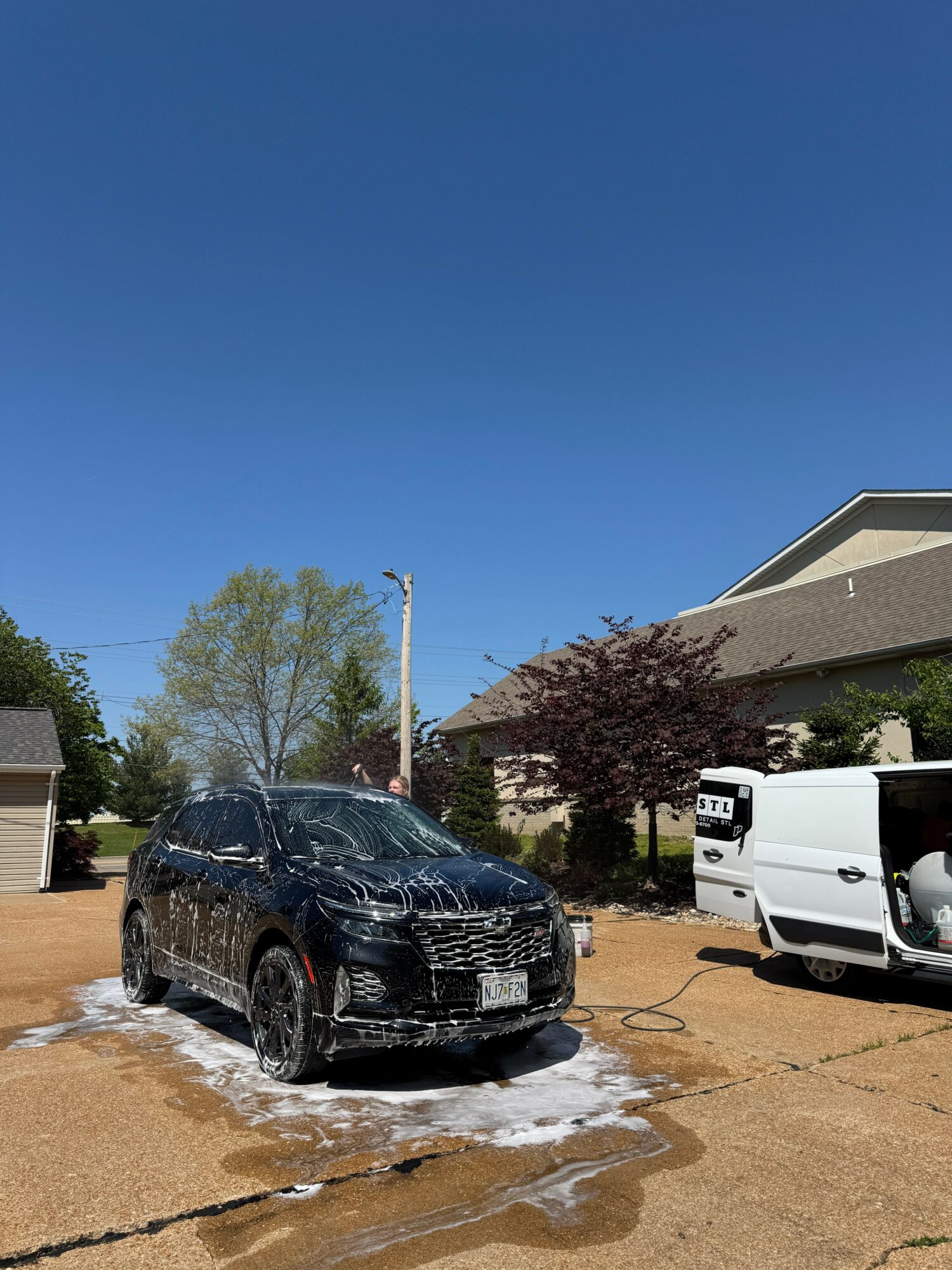 Black car parked on wet concrete with trees and buildings in background under clear blue sky.