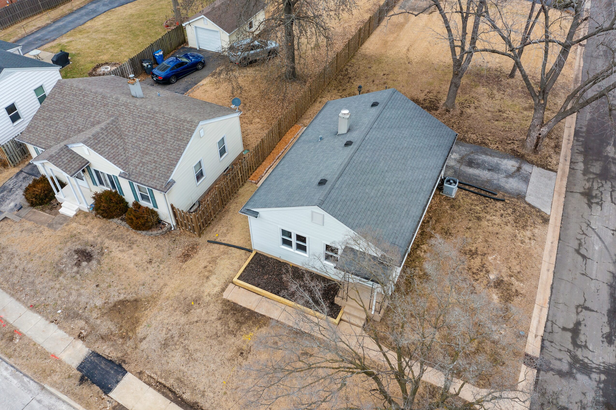 Aerial view of a house with a gray roof, backyard, and surrounding yard, with neighboring houses and trees nearby.
