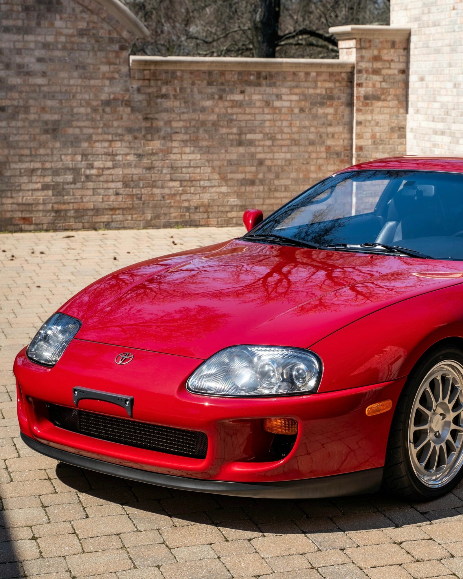 Red sports car parked on brick pavement with brick wall and tree in background.