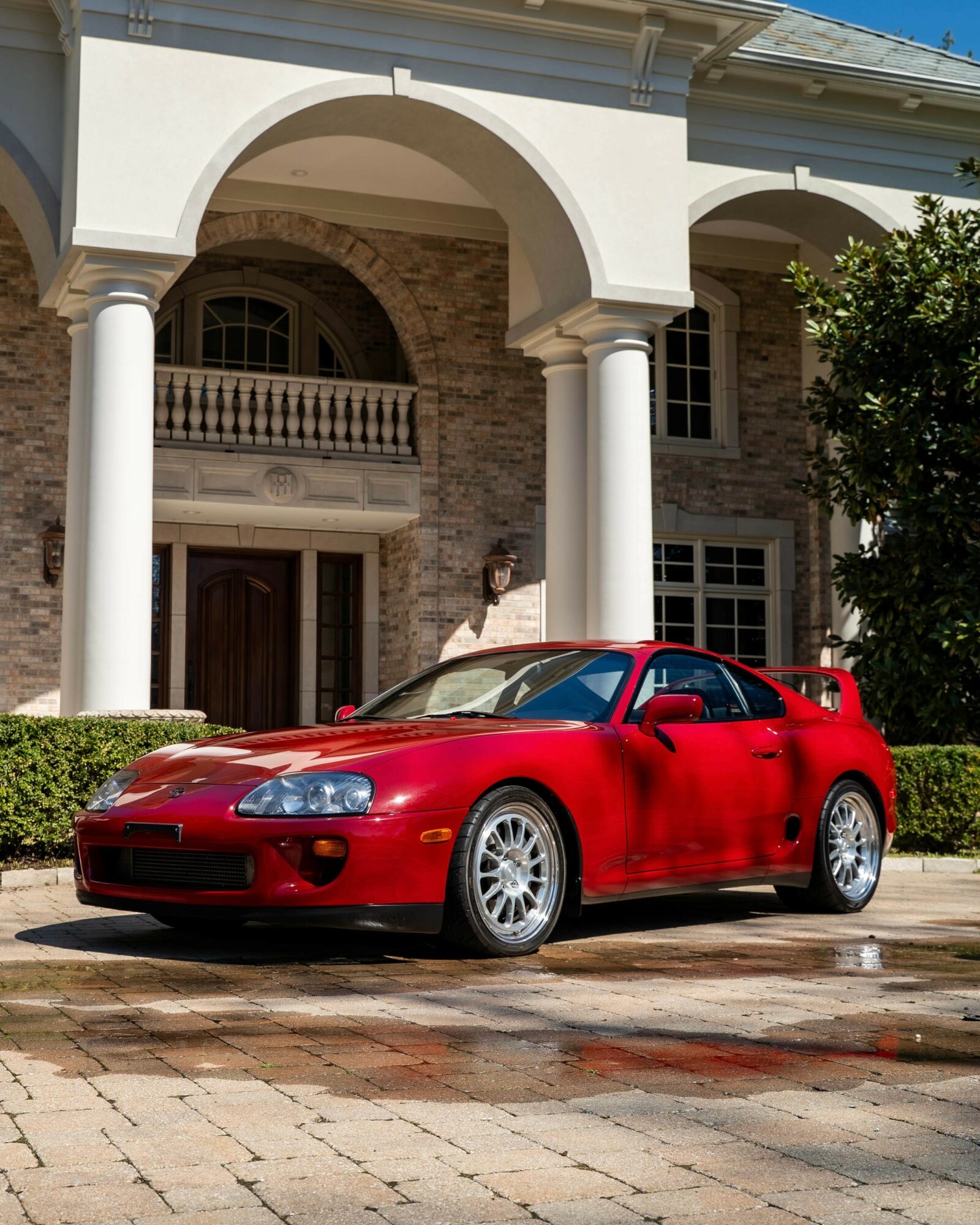 Red sports car parked in front of a large house with white columns and stone facade, sunny day.