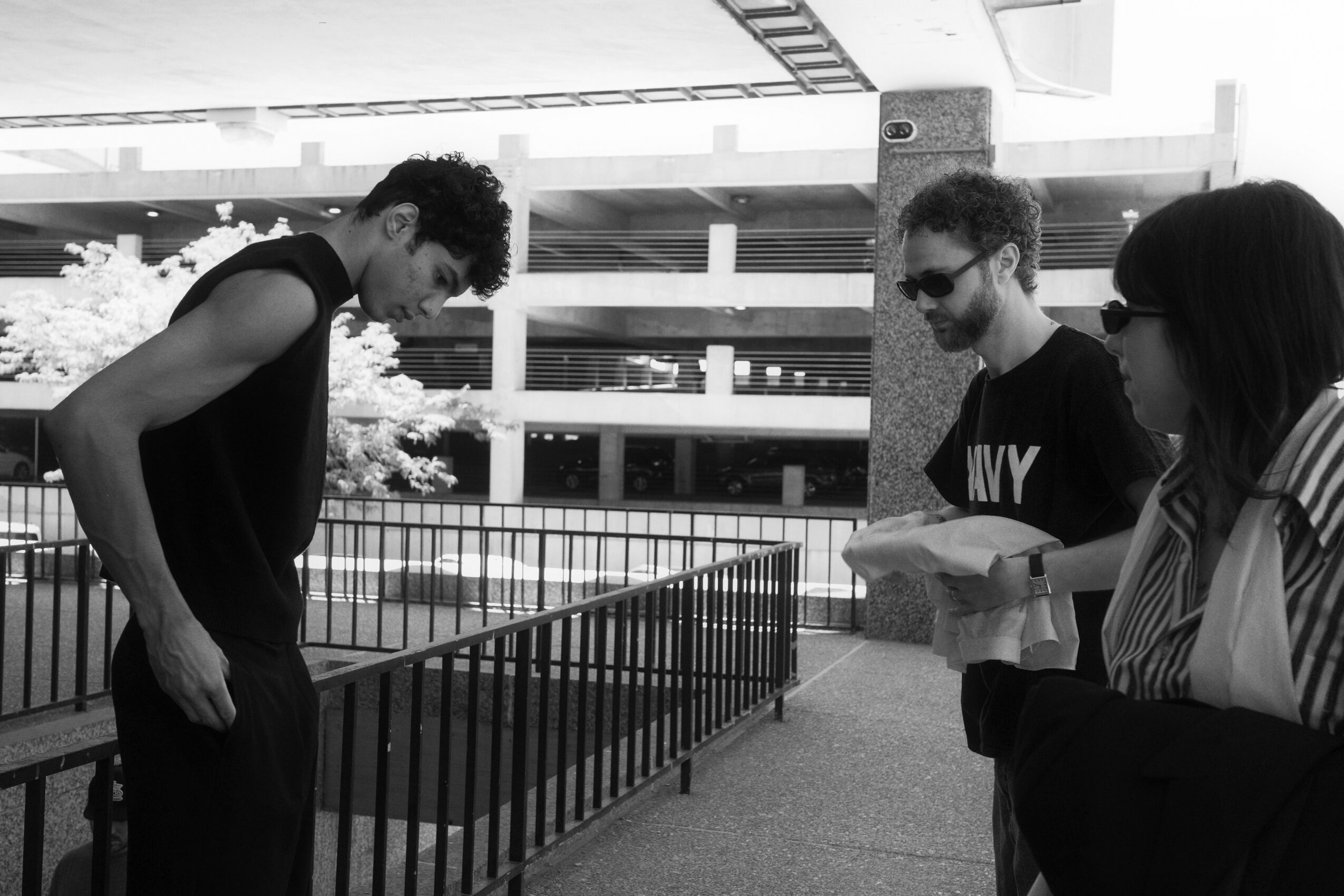 Three people standing outdoors near a railing, two men and one woman, engaged in conversation or reading, with a building in the background.