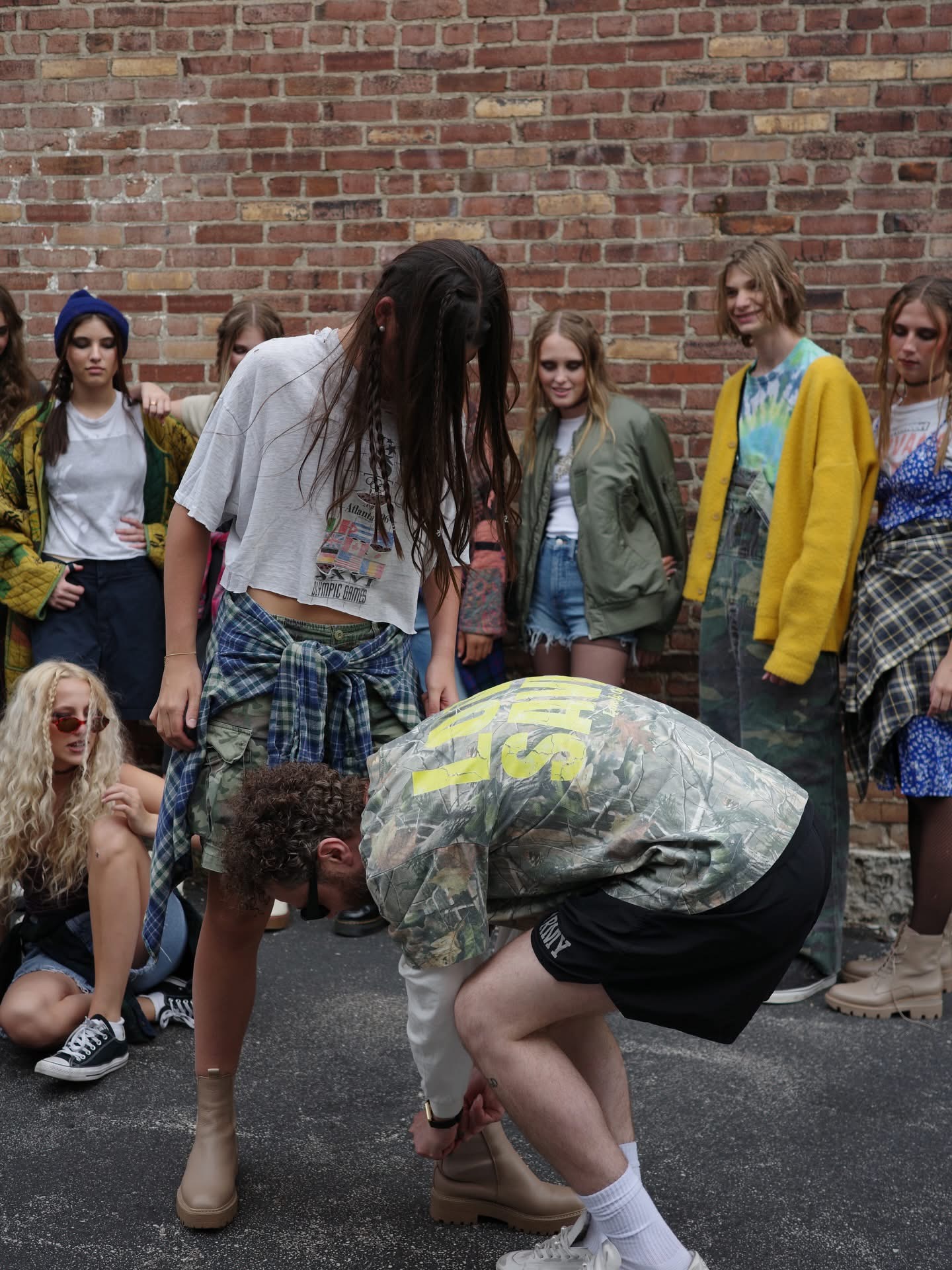 Group of young people standing against a brick wall, with one person crouching down in front. Casual clothing and varied expressions.