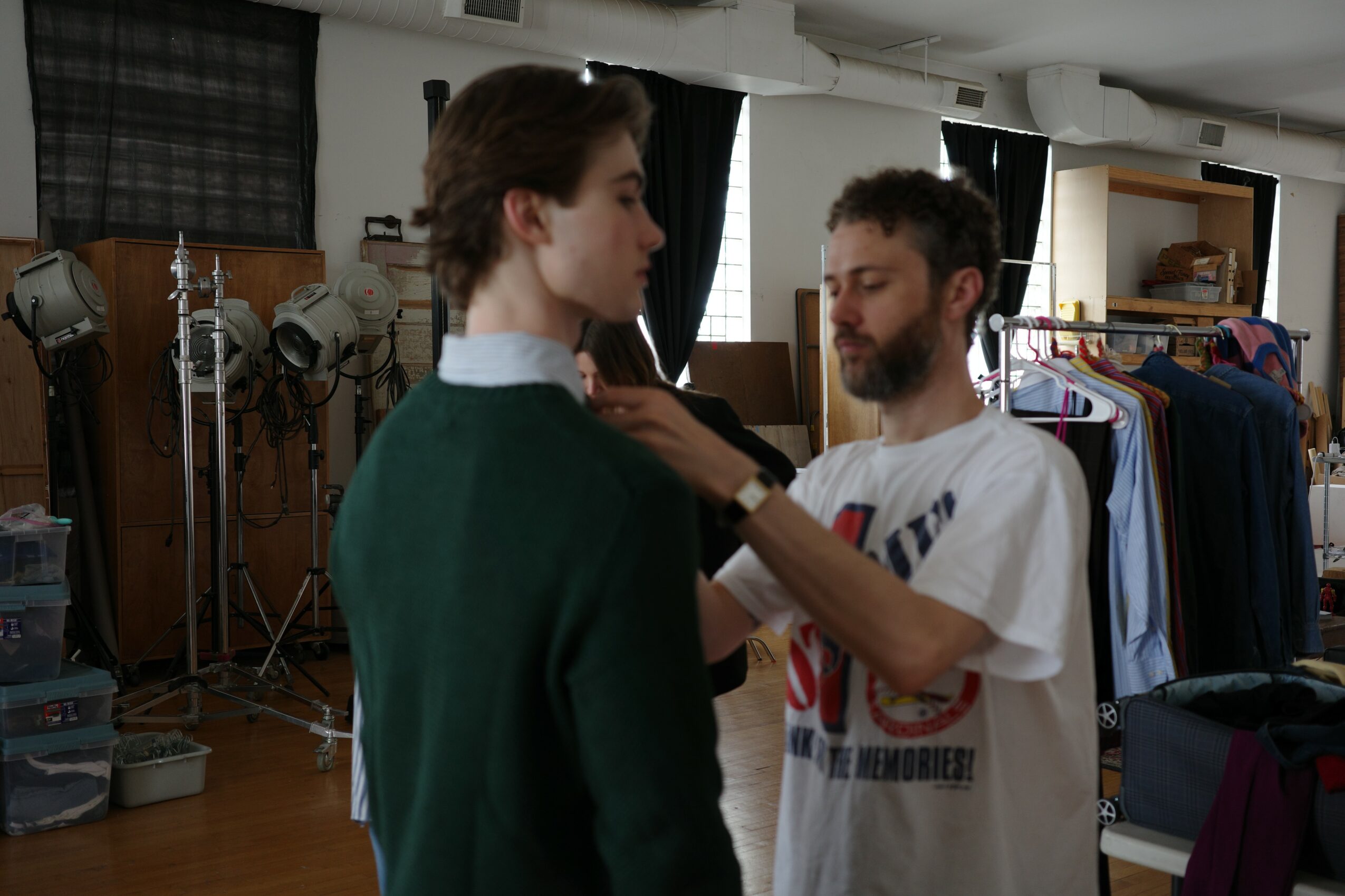 Two men in a room, one adjusting the other's collar, with clothing racks and equipment in the background.