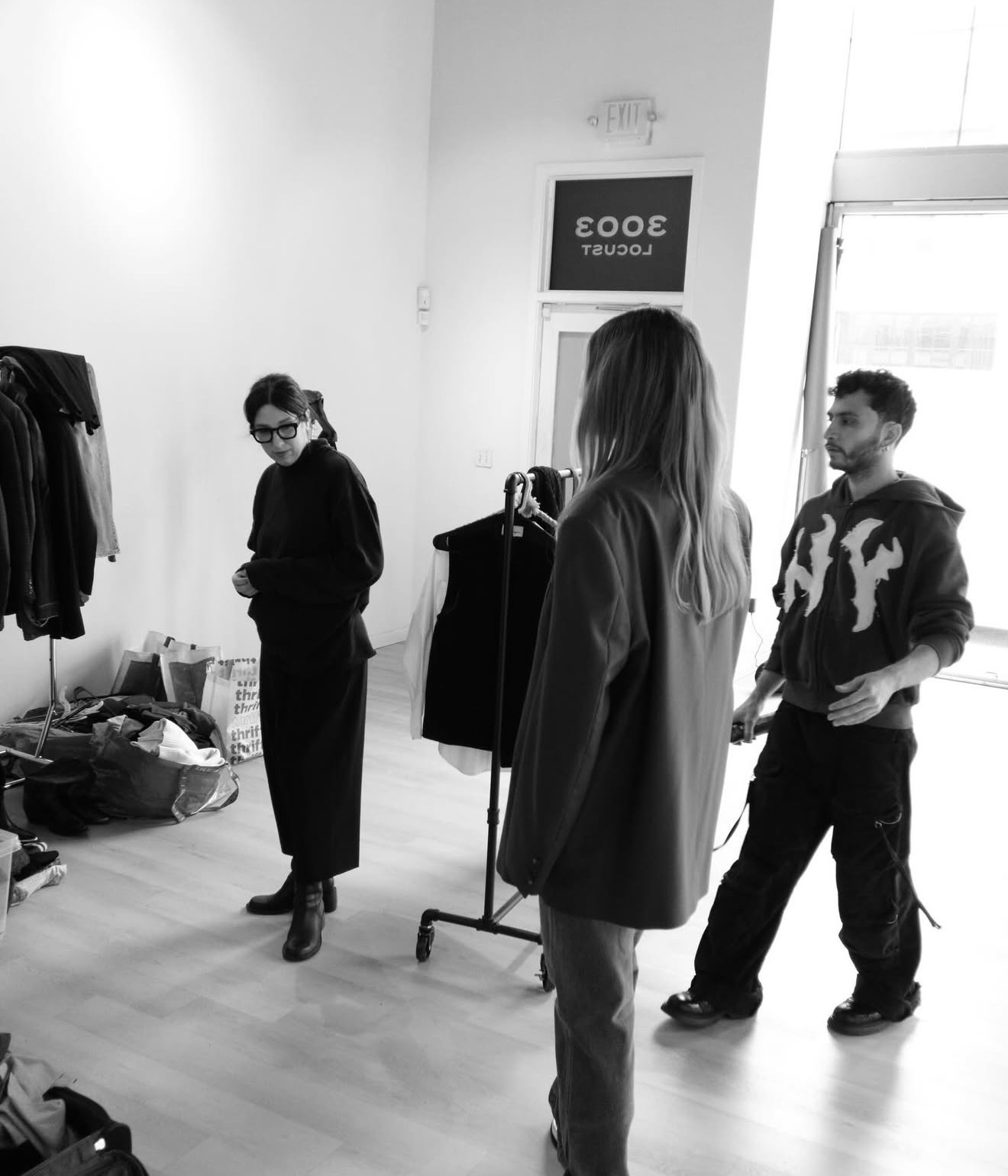 Three people in a room with clothing racks and a sign on the wall, one person facing away, two facing each other.