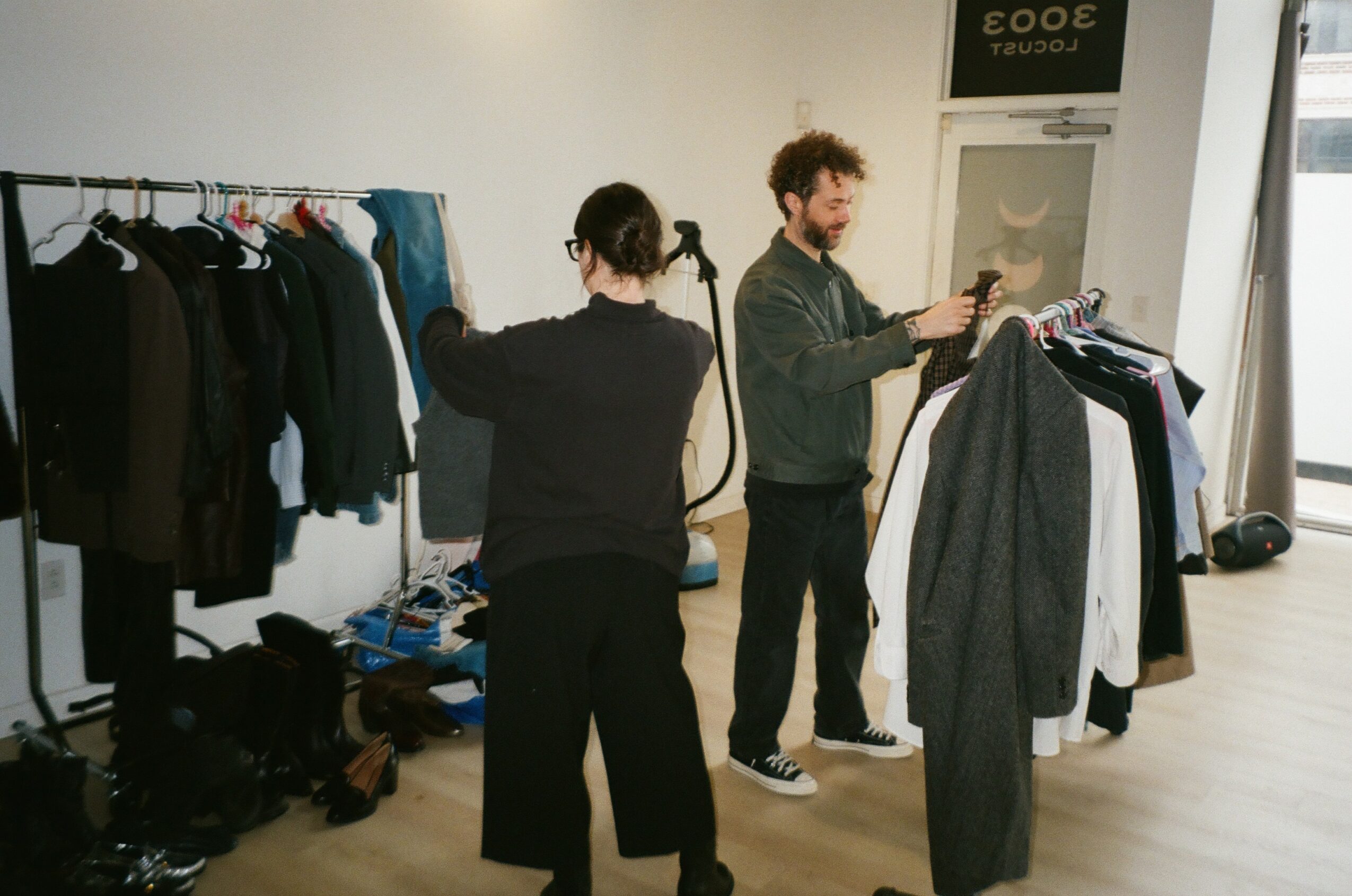 Two people in a clothing store, one browsing clothes on a rack, the other looking at a mirror, with clothing and shoes around.