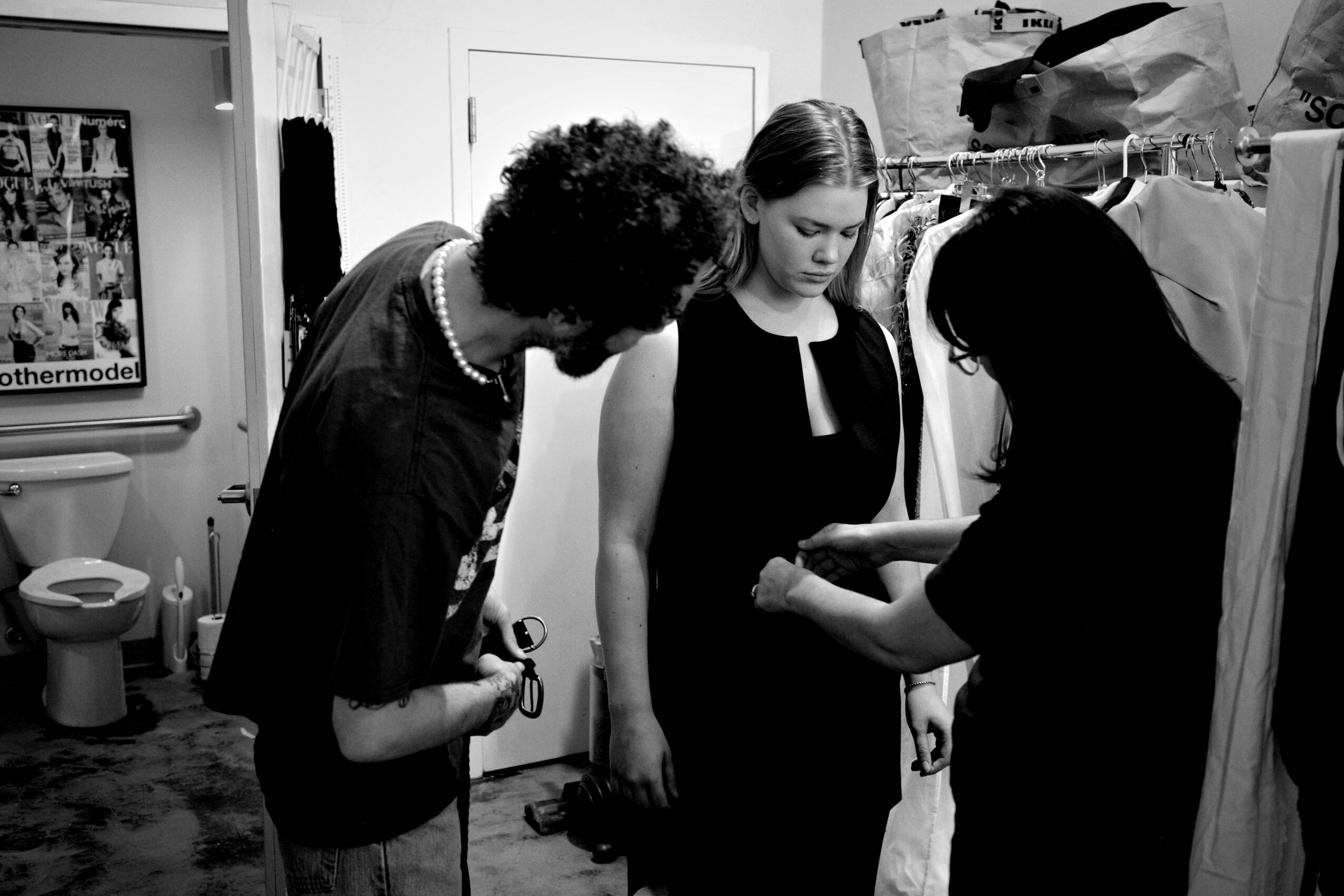 Three women in a room, one adjusting the other's sleeve, with a bathroom visible in the background.