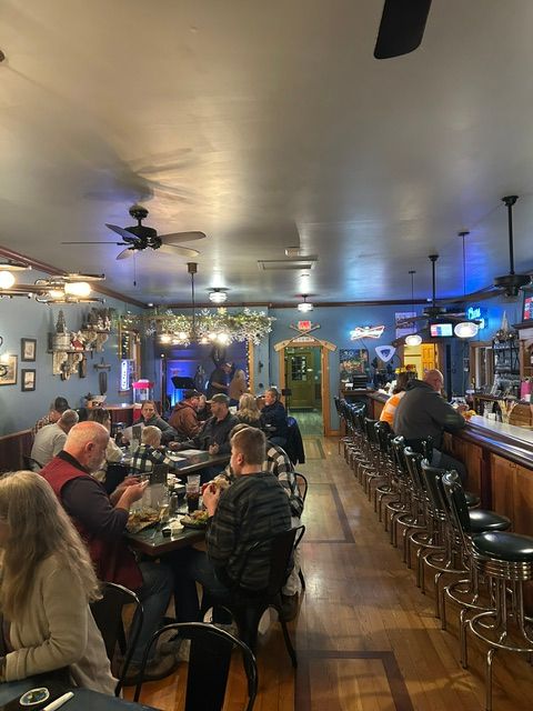 People sitting at tables in a restaurant or bar, with a bar counter and bottles in the background.