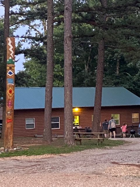 People gather outside a building with a blue roof, trees in the background, and a wooden utility pole in the foreground.