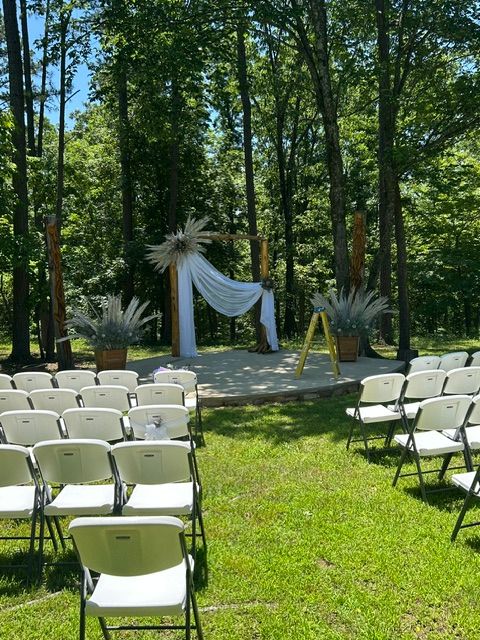 Outdoor scene with white chairs arranged in rows and decorated with white fabric and feathers, surrounded by trees and greenery.