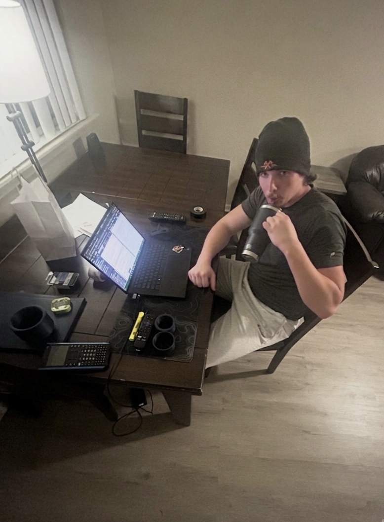 Person sitting at a desk with a laptop, wearing a beanie, in a room with a table, chair, and lamp.