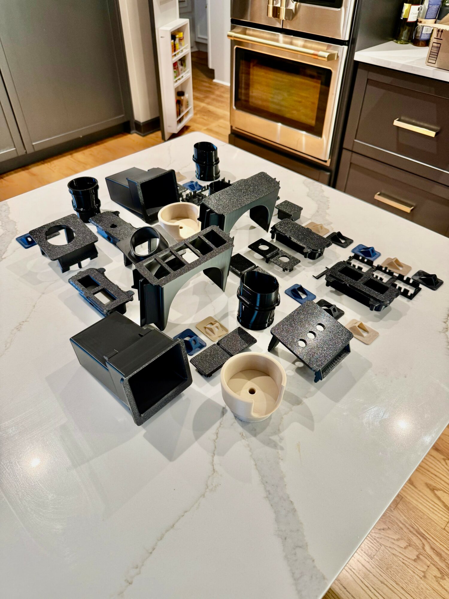 Various black electronic and mechanical components arranged on a white marble kitchen countertop with wooden flooring and appliances in background.
