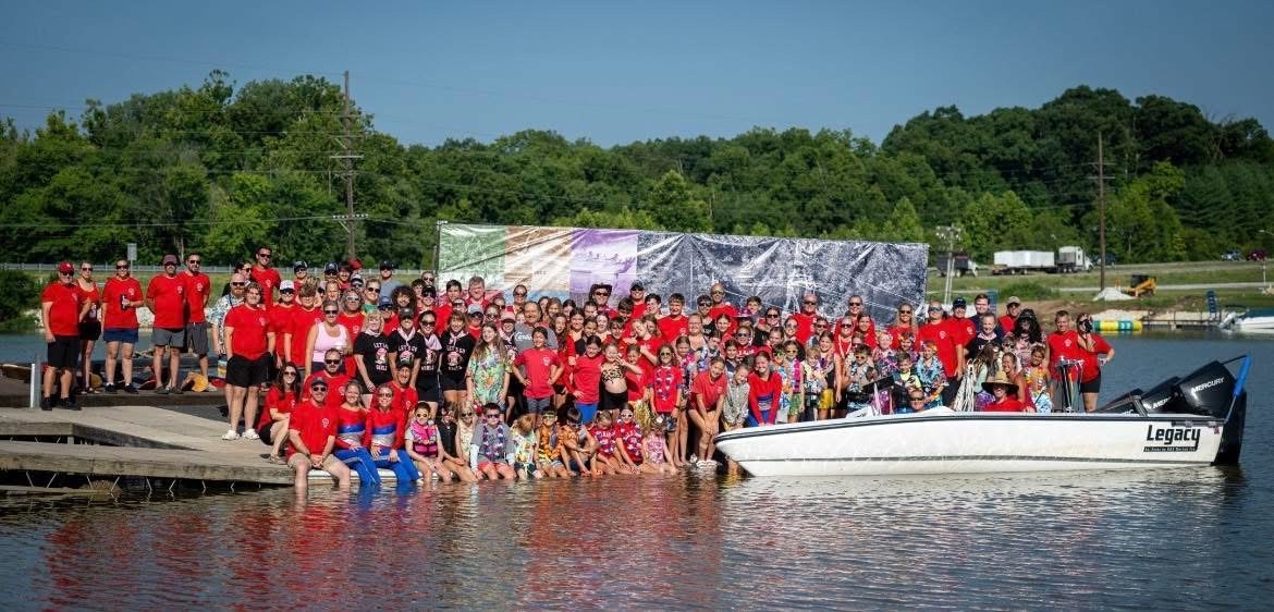 Large group of people in red shirts gathered by a dock with a boat on water, trees, and a blue sky in background.