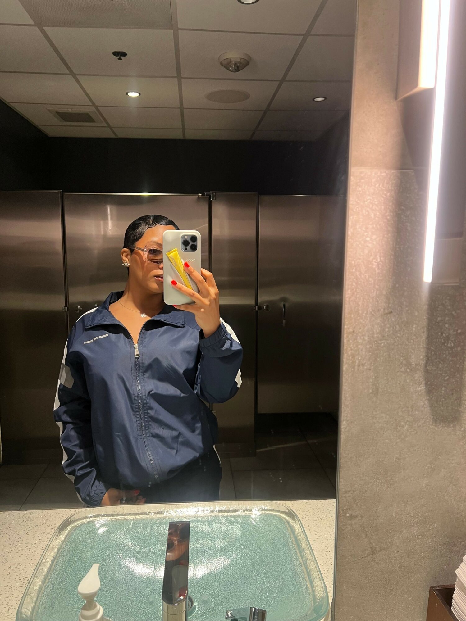 Person taking a mirror selfie in a bathroom, holding a phone with a yellow case, wearing a dark jacket, with a sink and mirror visible.