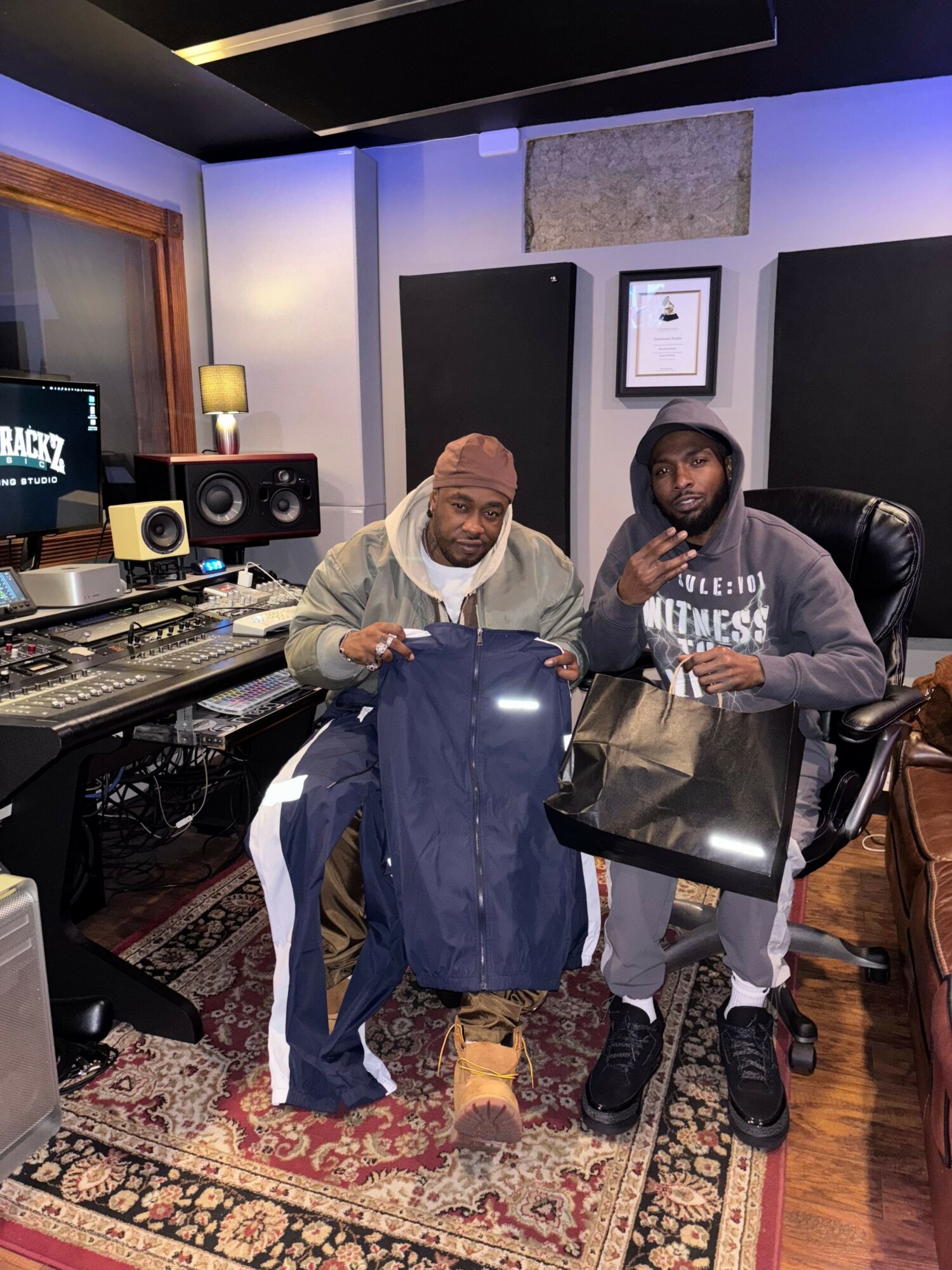 Two people in a music studio, one holding a shopping bag, the other taking a selfie, with audio equipment and a rug.