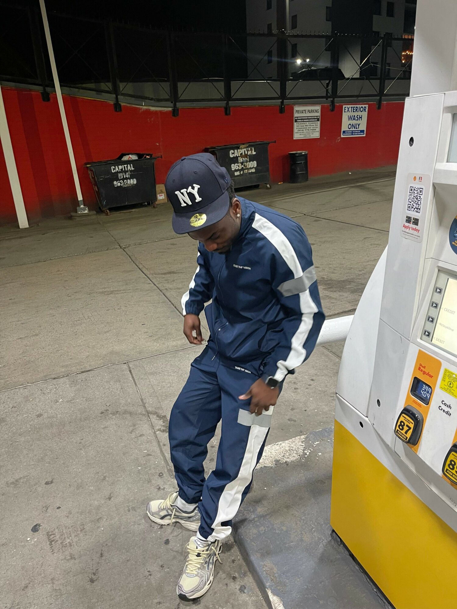 Person in blue tracksuit and cap at gas station, standing next to fuel pump, holding a phone, facing sideways.